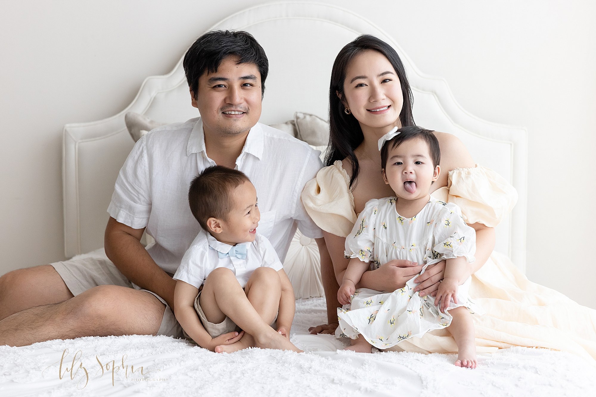  Fun family photo of an Asian father sitting on a bed with his toddler son in front of him watching his one year old sister who is sticking out her tongue as she sits on her mother’s lap next to them taken next to a window streaming natural light int