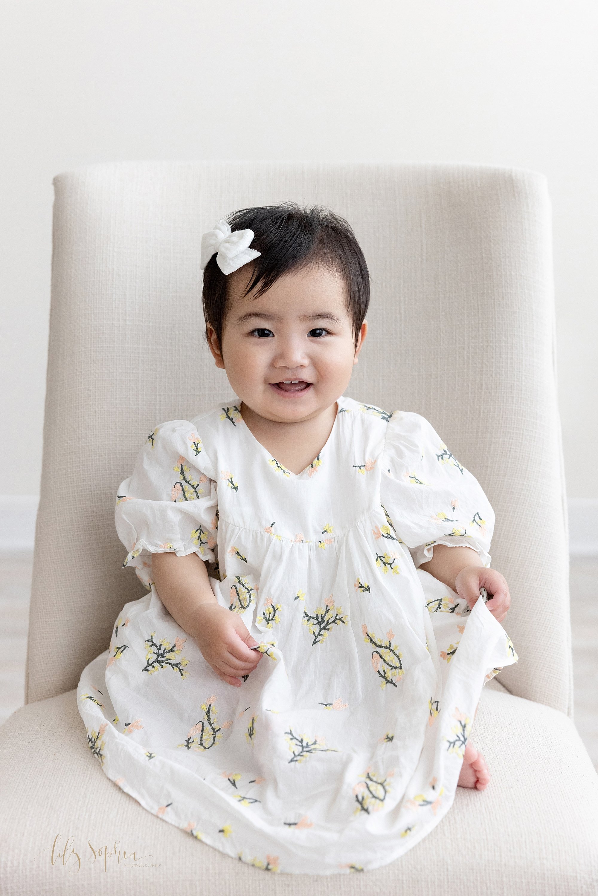  First birthday portrait of a proud one year old Asian girl as she sits smiling in an upholstered chair showing her tiny teeth taken using natural light in a photography studio located near Brookhaven in Atlanta. 
