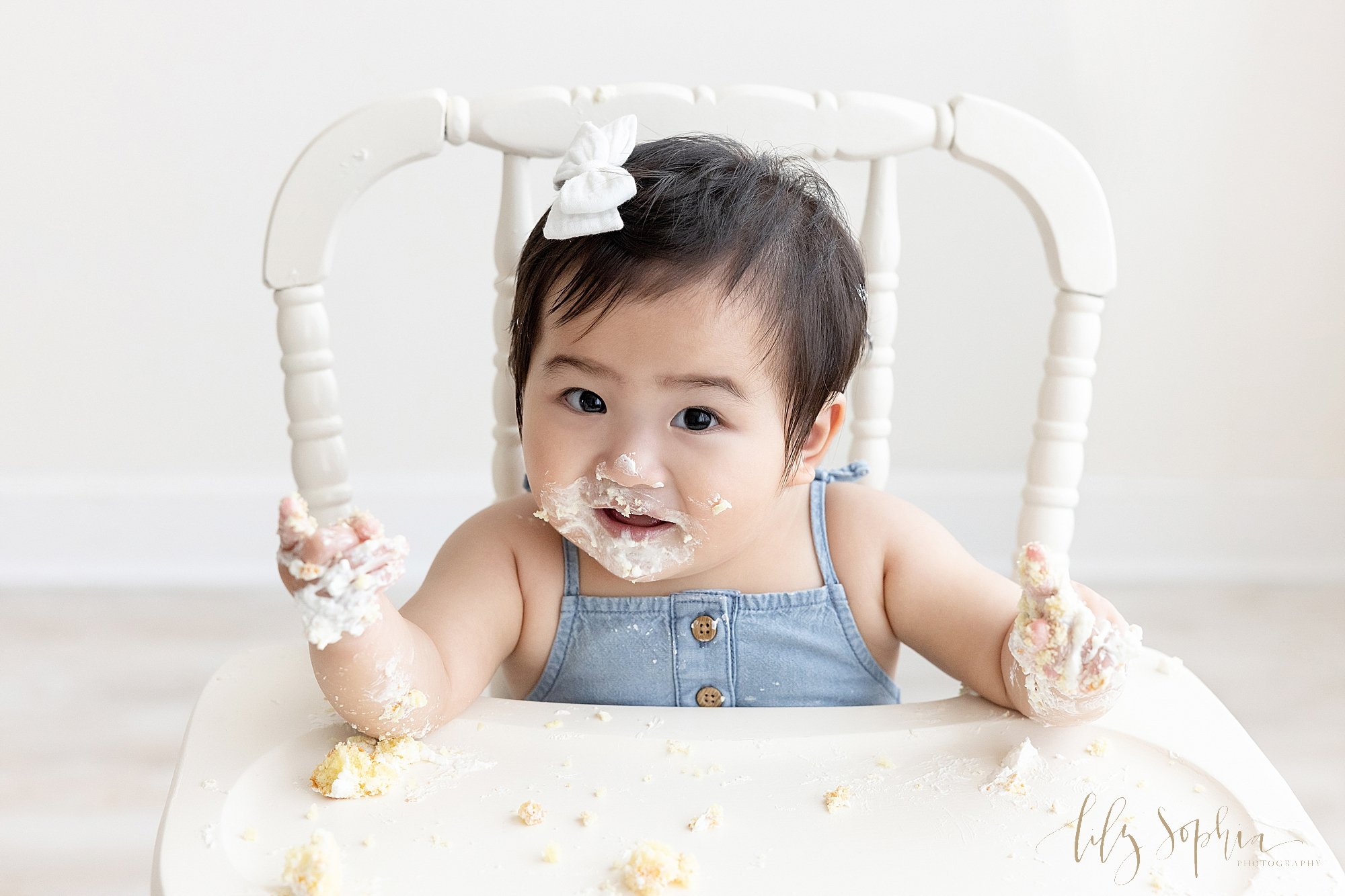  First birthday smash cake picture of a one year old Asian girl as she sits in an antique high chair with icing covering her face and hands with nothing but crumbs left on the tray taken near Roswell in Atlanta in a photography studio that uses natur