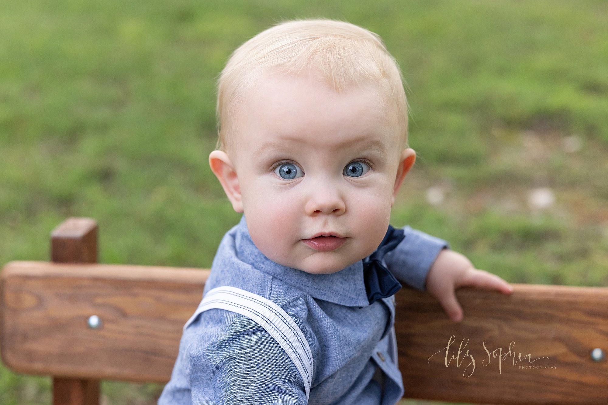  Close-up photo of a blue eyed baby boy as he sits on a wooden bench at sunset in a park near Atlanta. 