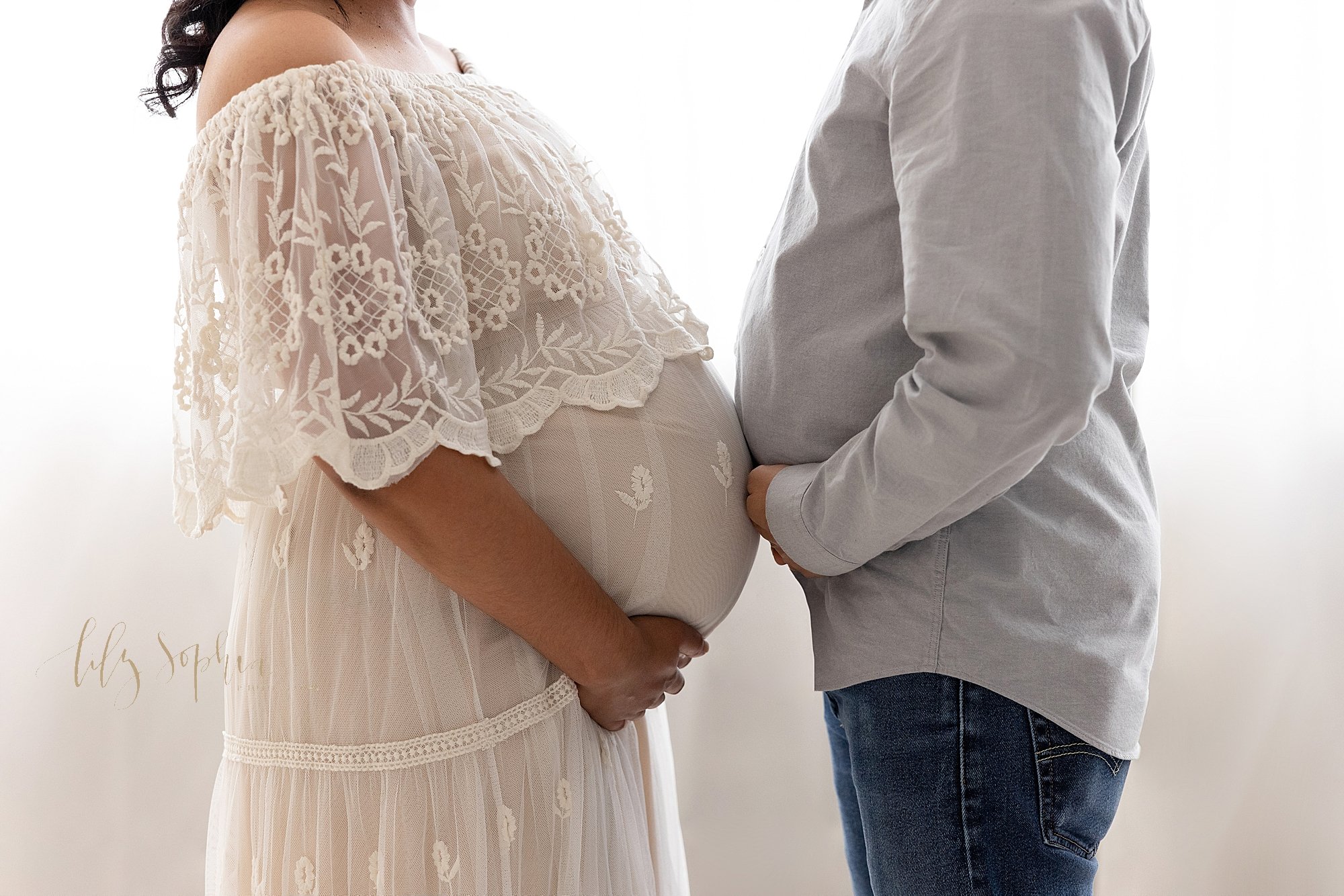  Maternity photo of a pregnant mother holding the base of her belly facing her husband that is also holding the base of his belly as their bellies tough while they stand in front of a window streaming natural light in a studio near Roswell in Atlanta