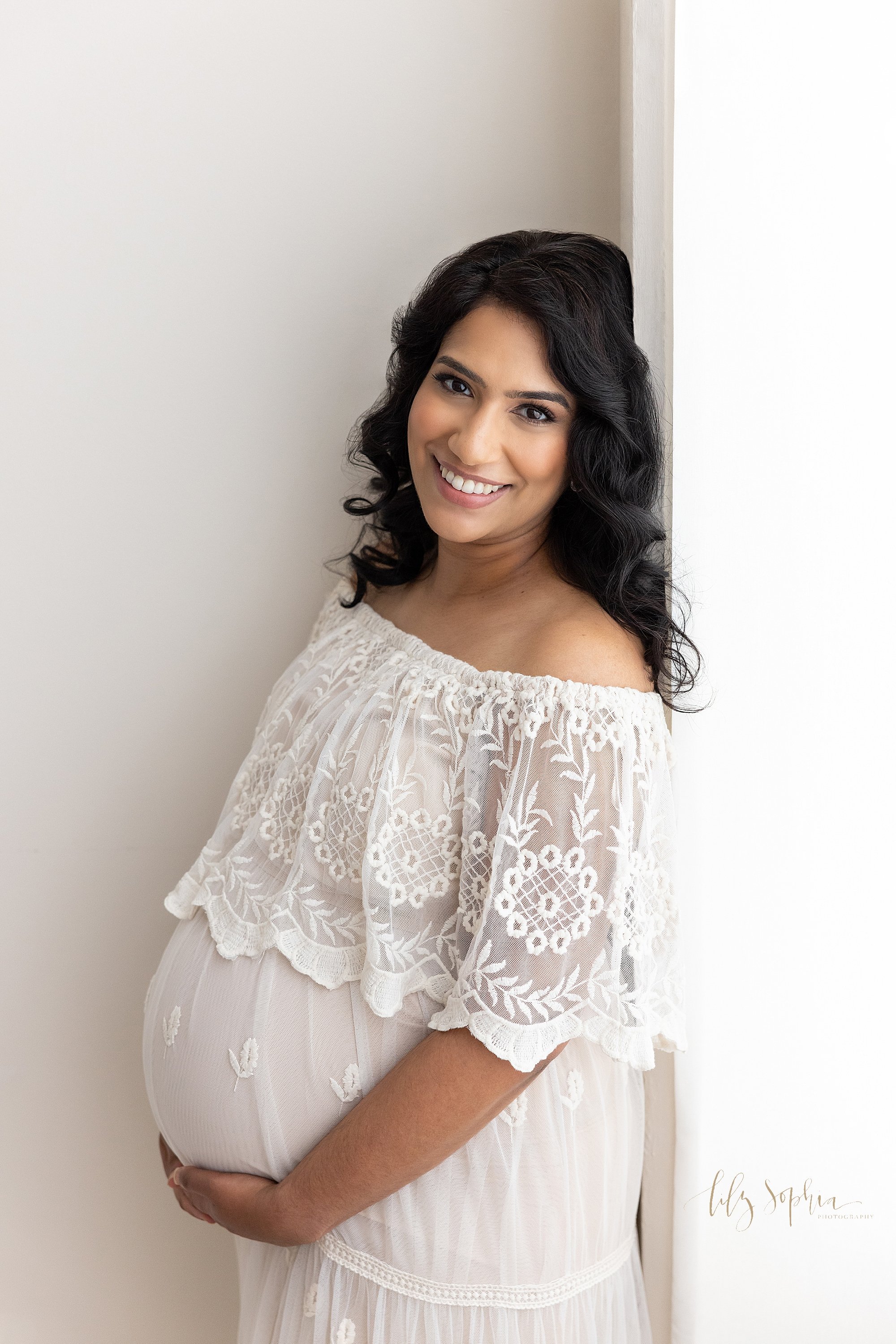  Maternity photo session of an expectant mother wearing an off the shoulder embroidered bodice flowing gown standing with her back against a window streaming natural light as she holds the base of her pregnant belly taken near Ansley Park in Atlanta 