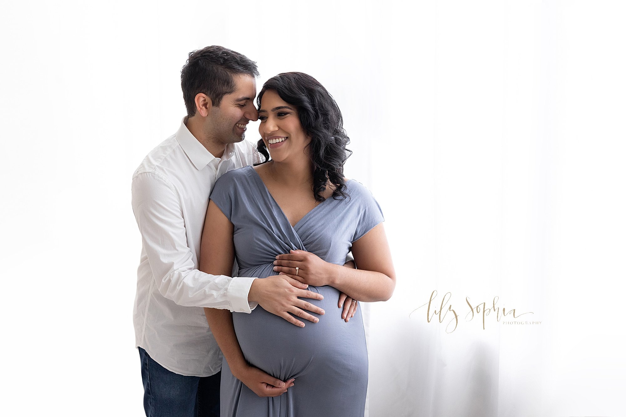  Maternity photo session with a pregnant mother standing in front of window streaming natural light with her hands framing her belly and her husband standing to her right side with his right hand on his child in utero while whispering into his wife’s