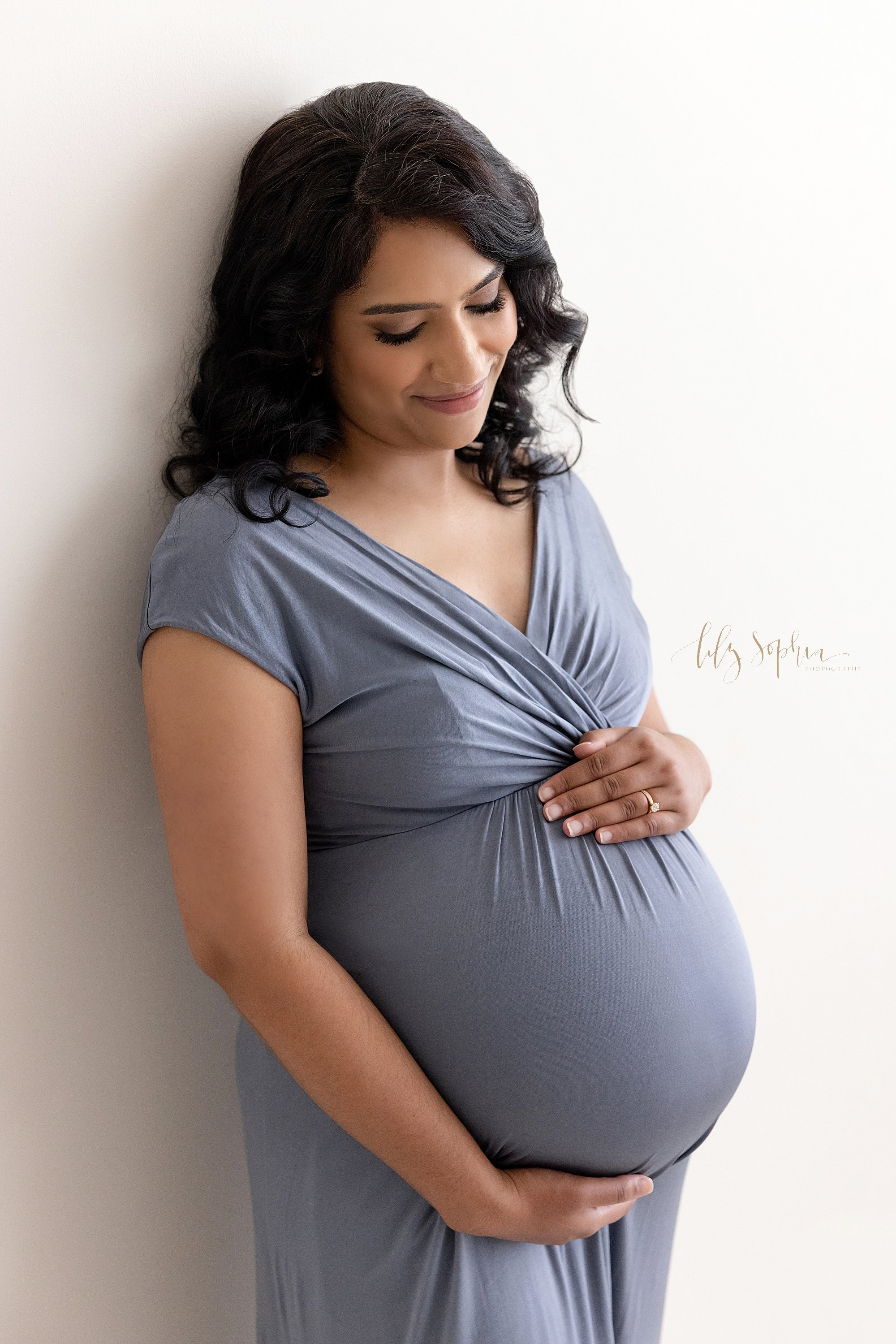  Maternity picture of a pregnant mother wearing a jersey knit V-neck gown as she stands against the wall of a photography studio framing her belly with her hands and contemplating the birth of her child taken near Brookhaven in Atlanta in natural lig