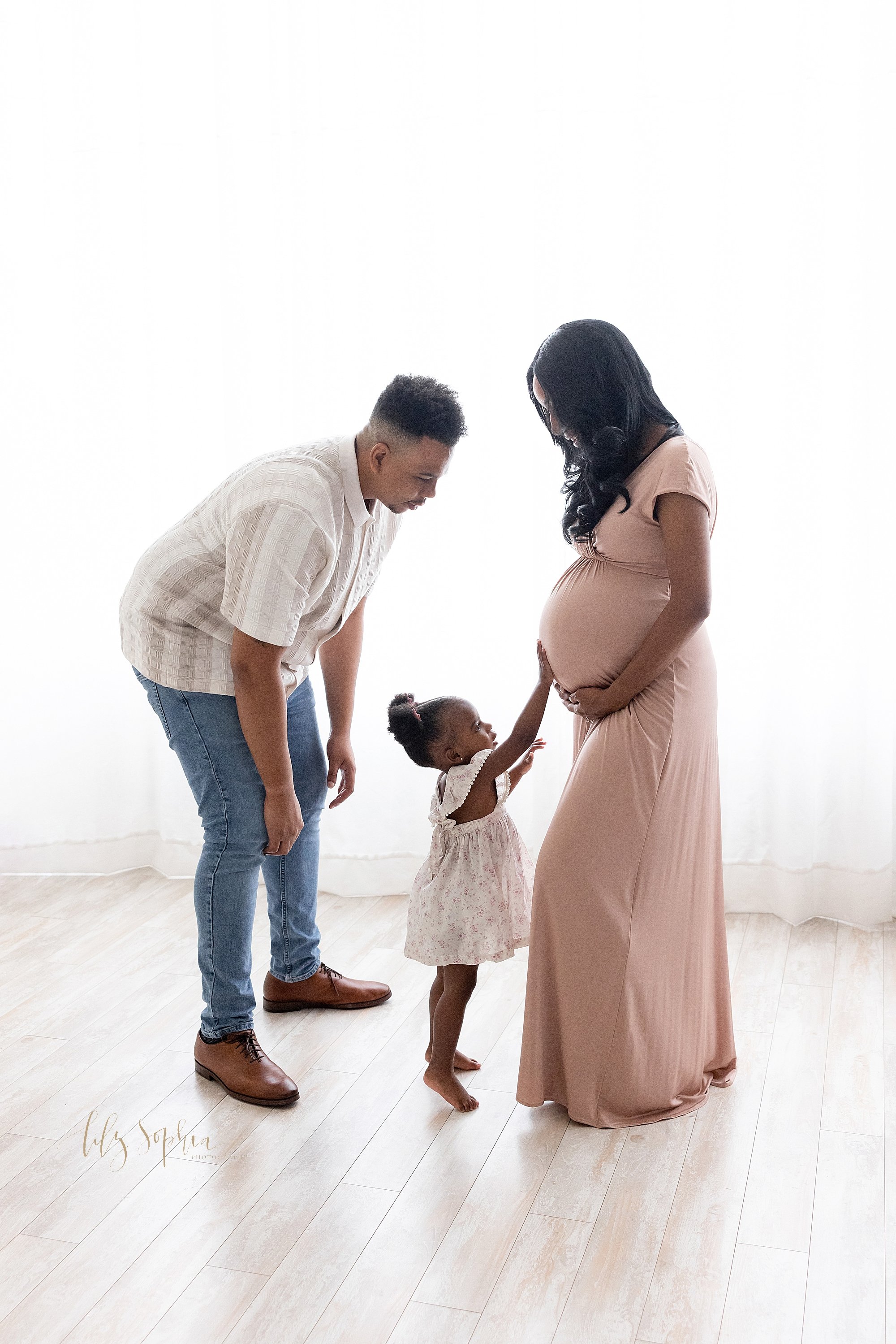  Maternity family photo session with a pregnant African-American mother holding the base of her belly as her toddler daughter places her hand on her sibling in utero and her husband faces her to guide their daughter taken in front of a window streami