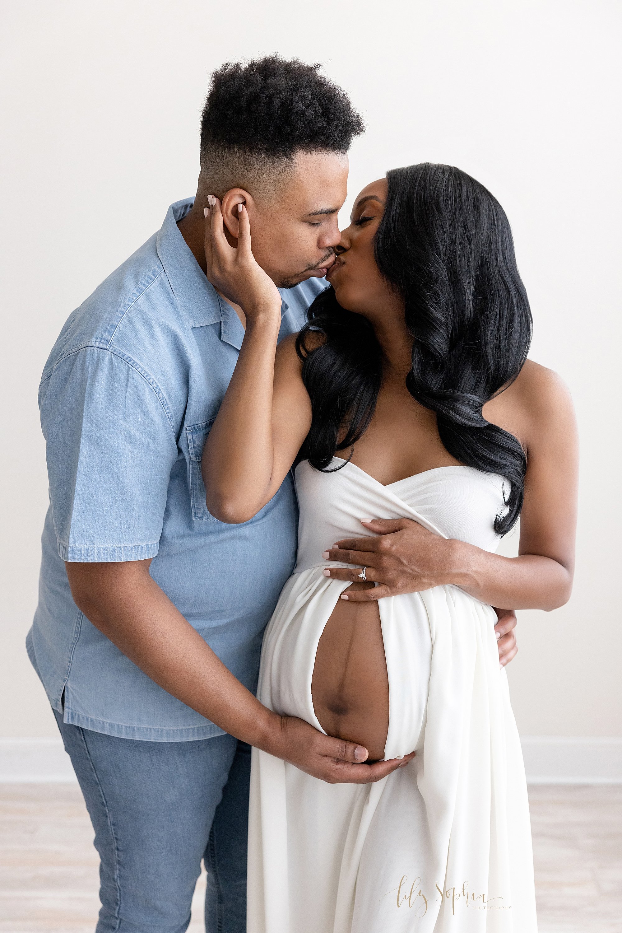  Maternity photo shoot with an African-American couple as they stand in a natural light studio with the pregnant mother baring her belly as she places her left hand on top of her belly and her husband places his right hand at the base of her belly an