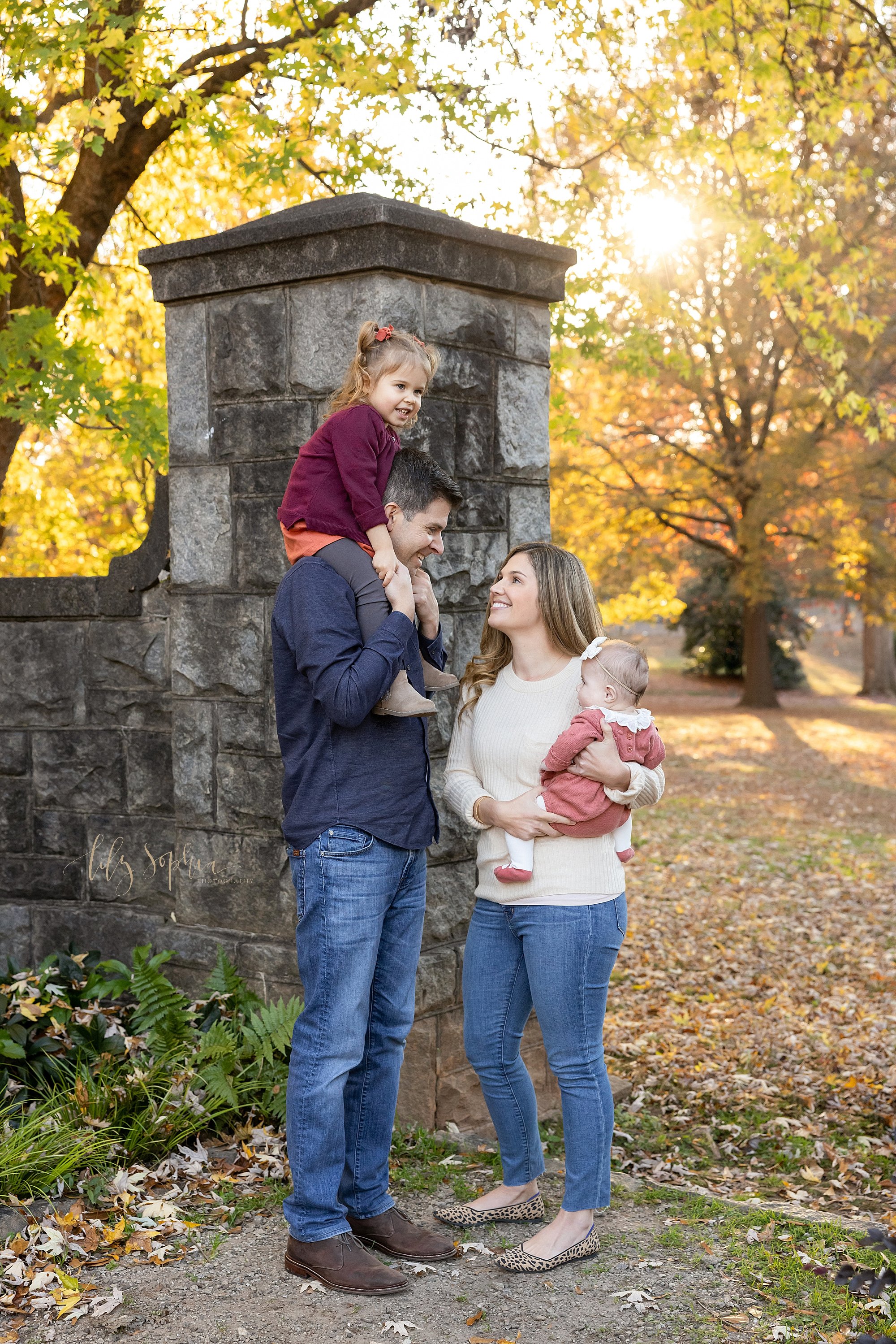  Fun family portrait taken at sunset in front of a stone entranceway at a park near Atlanta with a father carrying his toddler daughter on his shoulders as his wife holds their baby girl on her right hip as the two of them talk taken during autumn. 
