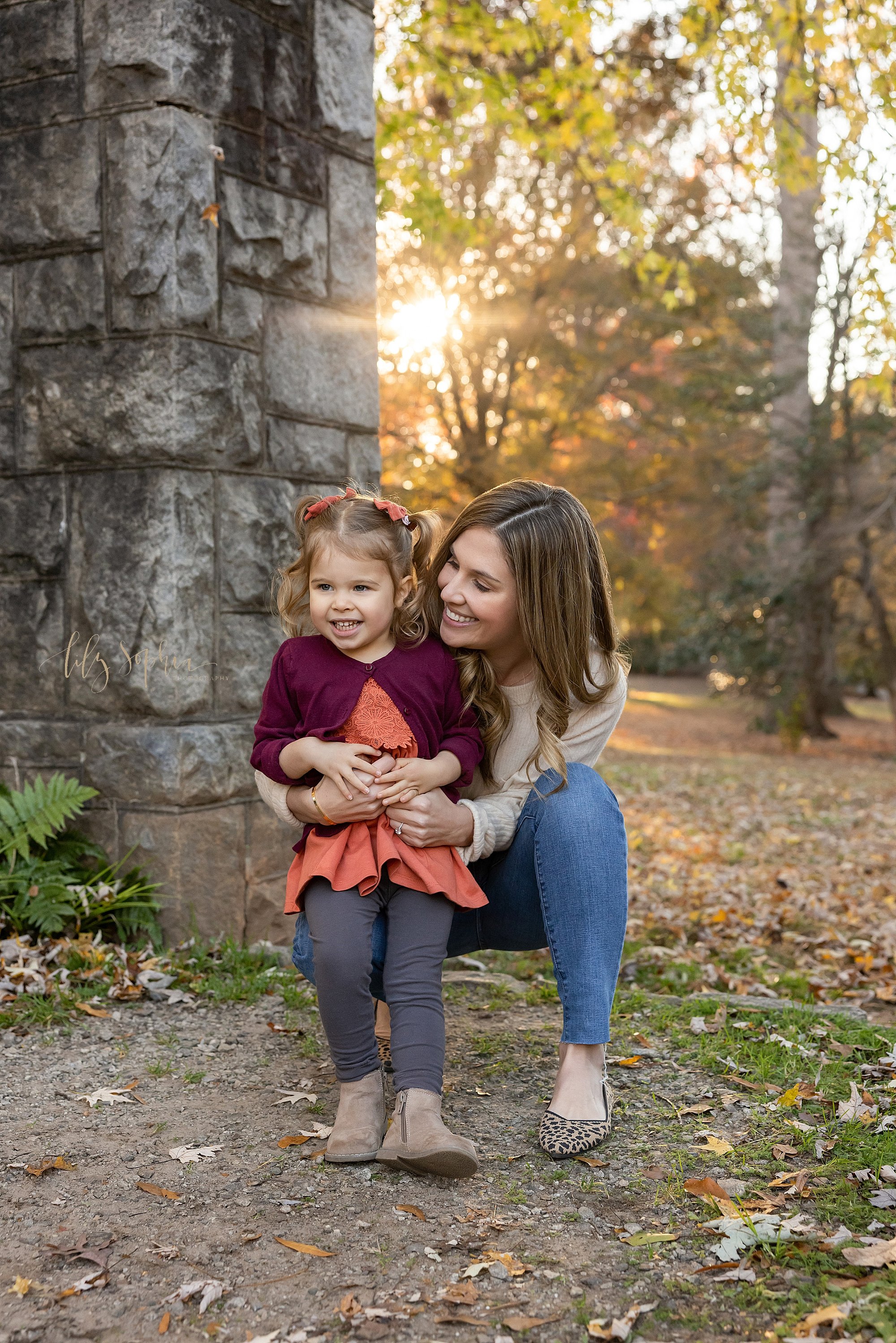  Family picture of a mother holding her toddler daughter on her right knee as she squats next to a stone entranceway during autumn at sunset in an Atlanta park. 
