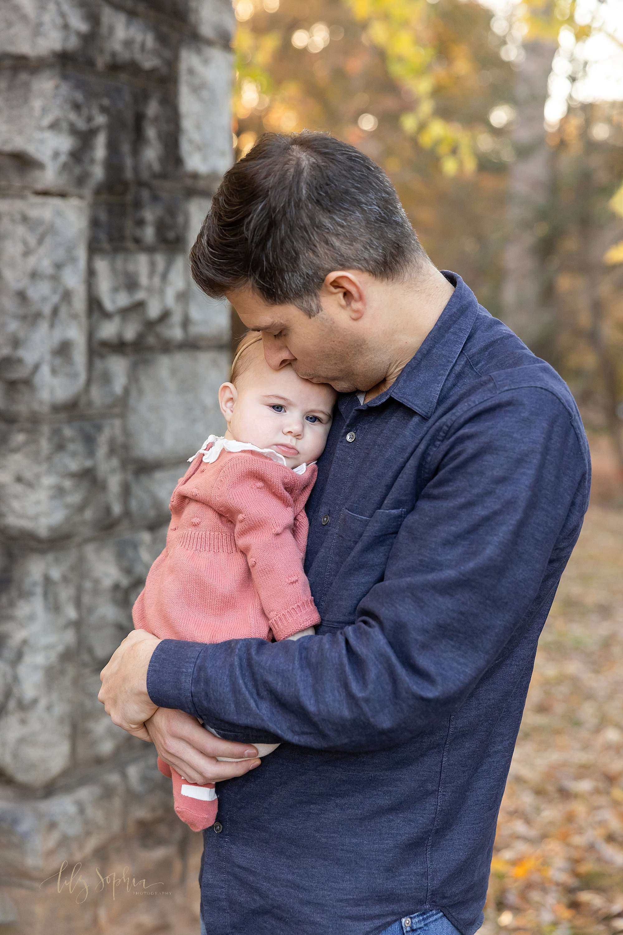  Sweet family photo of a father holding his baby girl against his chest as he kisses her forehead taken during autumn at sunset in a park near Atlanta in a stone entranceway. 