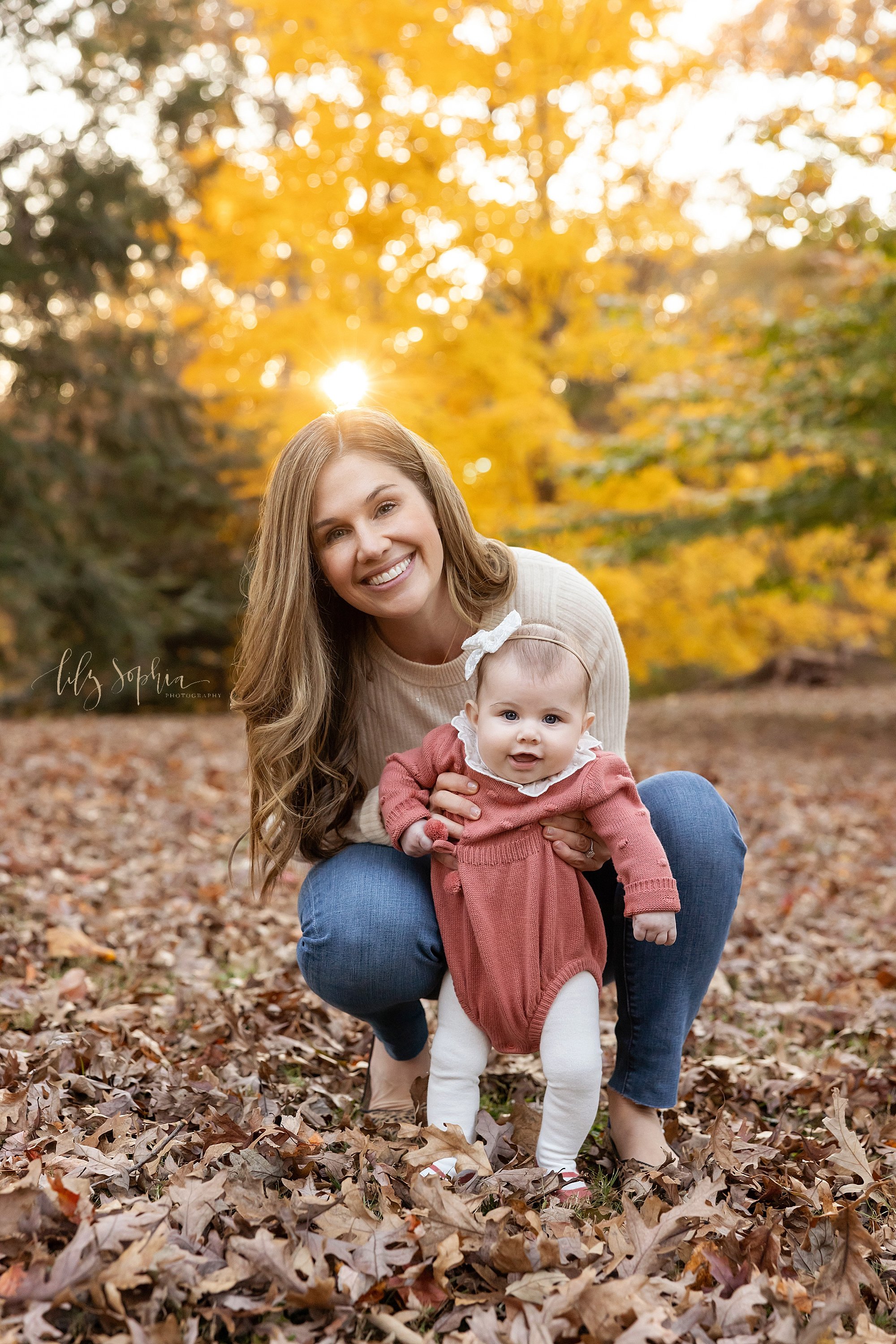  Family photo of a mother squatting in a park near Atlanta, Georgia holding her baby girl between her knees to support her as she stands amongst the leaves during autumn at sunset. 