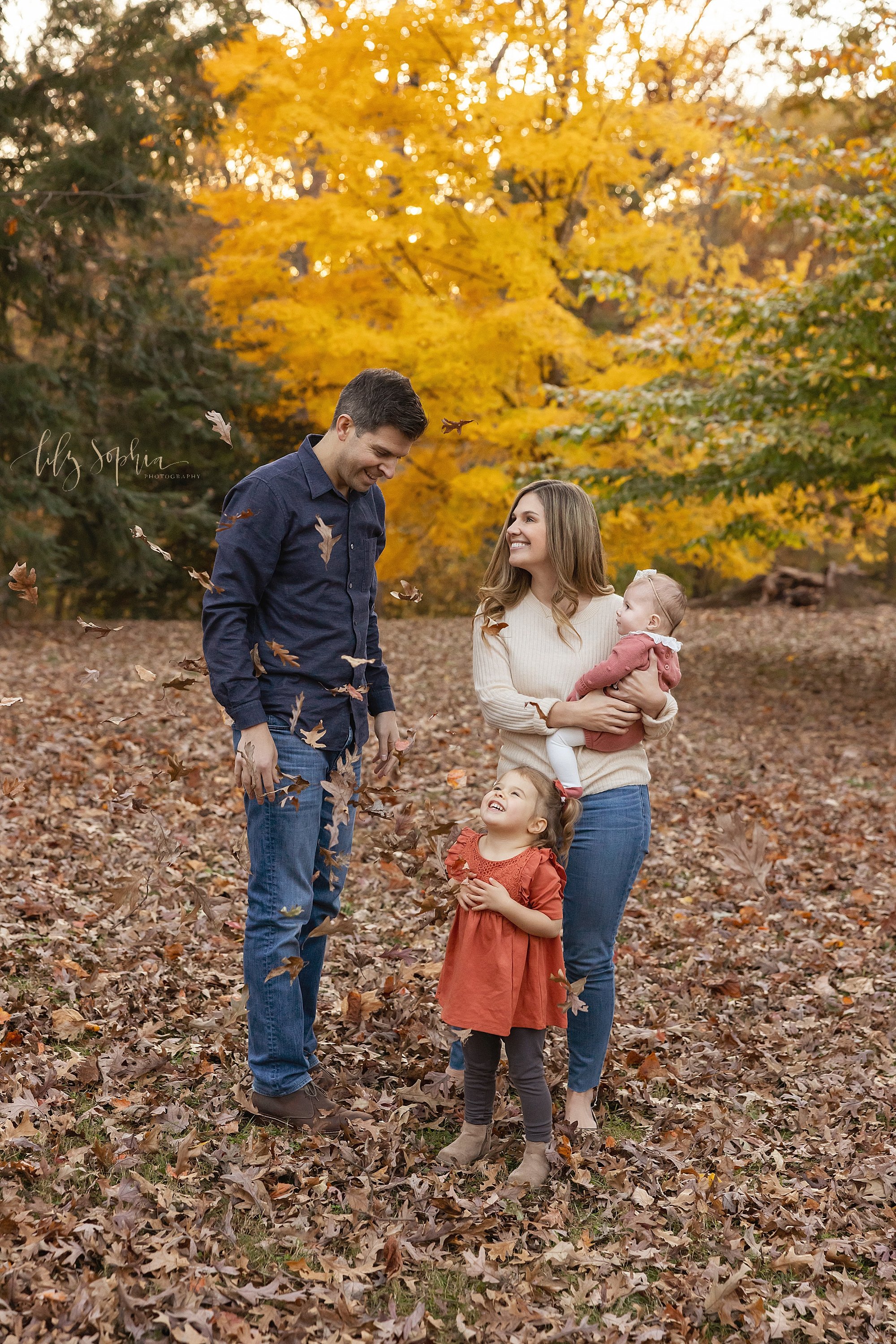  Family photo taken in a park near Atlanta during autumn with a father watching leaves he has tossed into the air fall while their toddler daughter looks up with delight and his wife stands to his left holding their baby daughter on her right hip adm