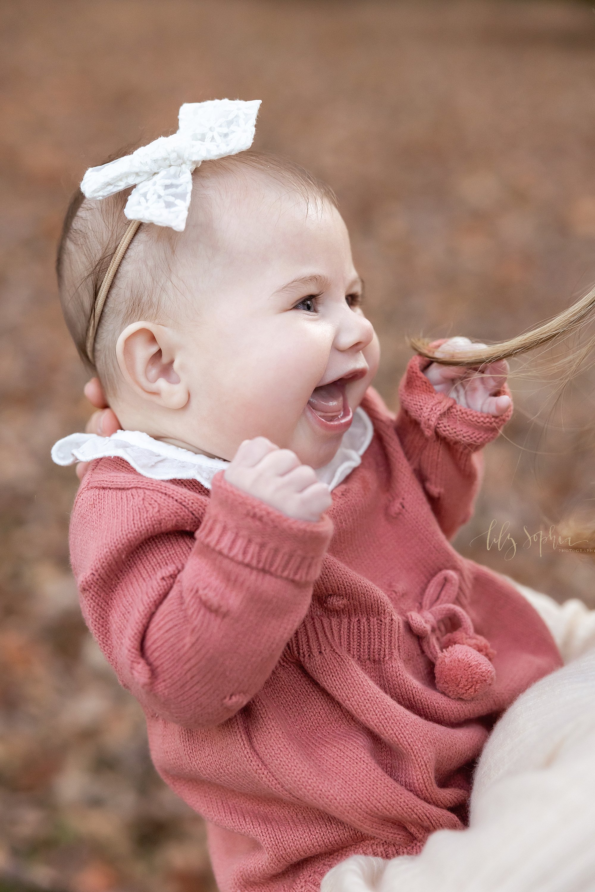 Close-up portrait of a happy baby girl as she is held in her mother’s arms and the baby grabs her hair taken during fall at sunset in a park near Atlanta. 