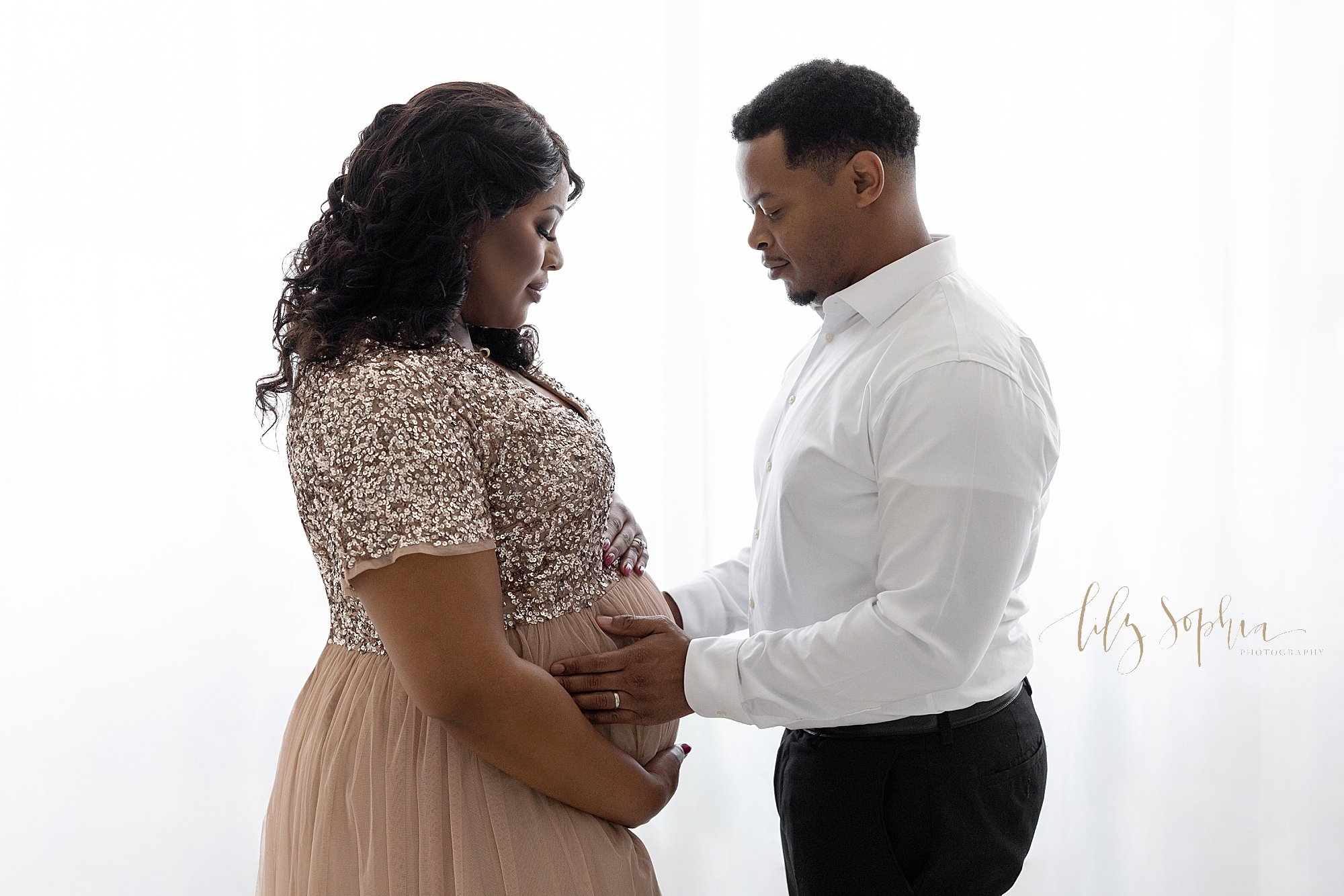  Maternity portrait of an African-American expectant couple facing one another as mom holds the base of her belly and dad places his hands on their child in utero as they contemplate the upcoming birth while they stand in front of a window streaming 