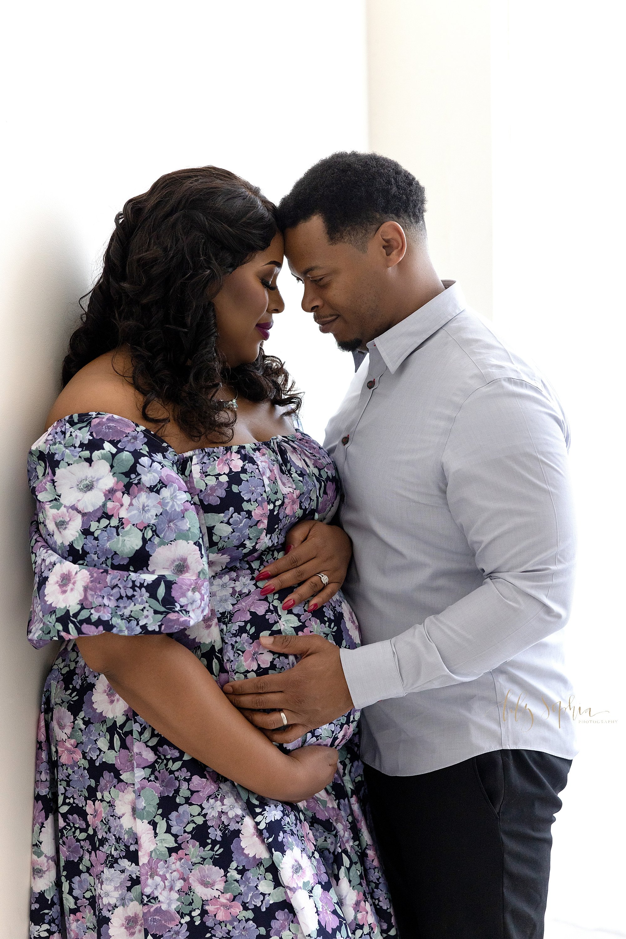  Maternity portrait of an African-American mother standing with her back against a wall and framing her belly as her husband faces her with his hands on their child in utero as the two of them places their foreheads together taken in a natural light 