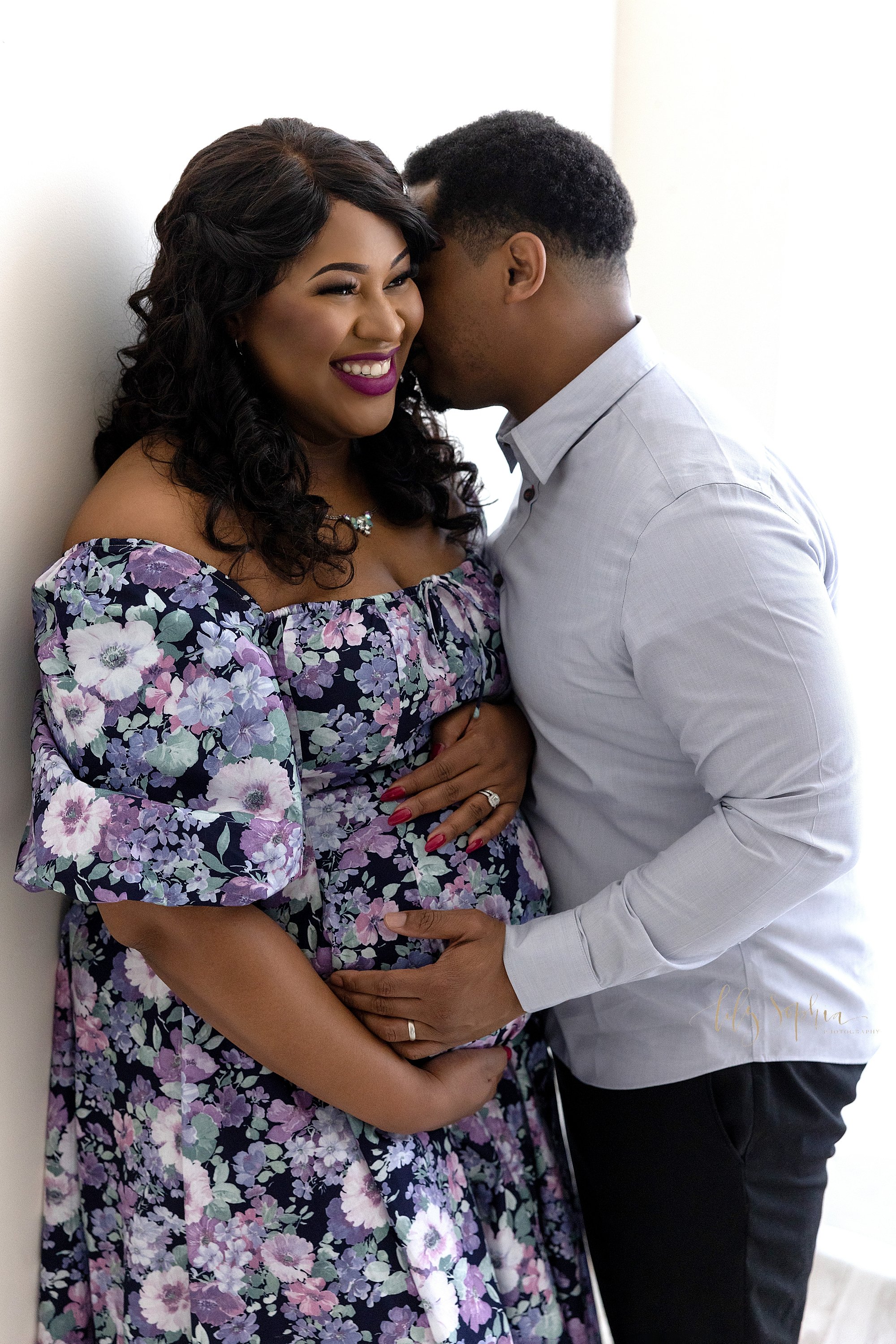  Maternity photo of a happy couple as the pregnant African-American woman frames her belly with her hands and her husband whispers in her left ear as he faces her and places his hand on their child in utero taken next to a window streaming natural li