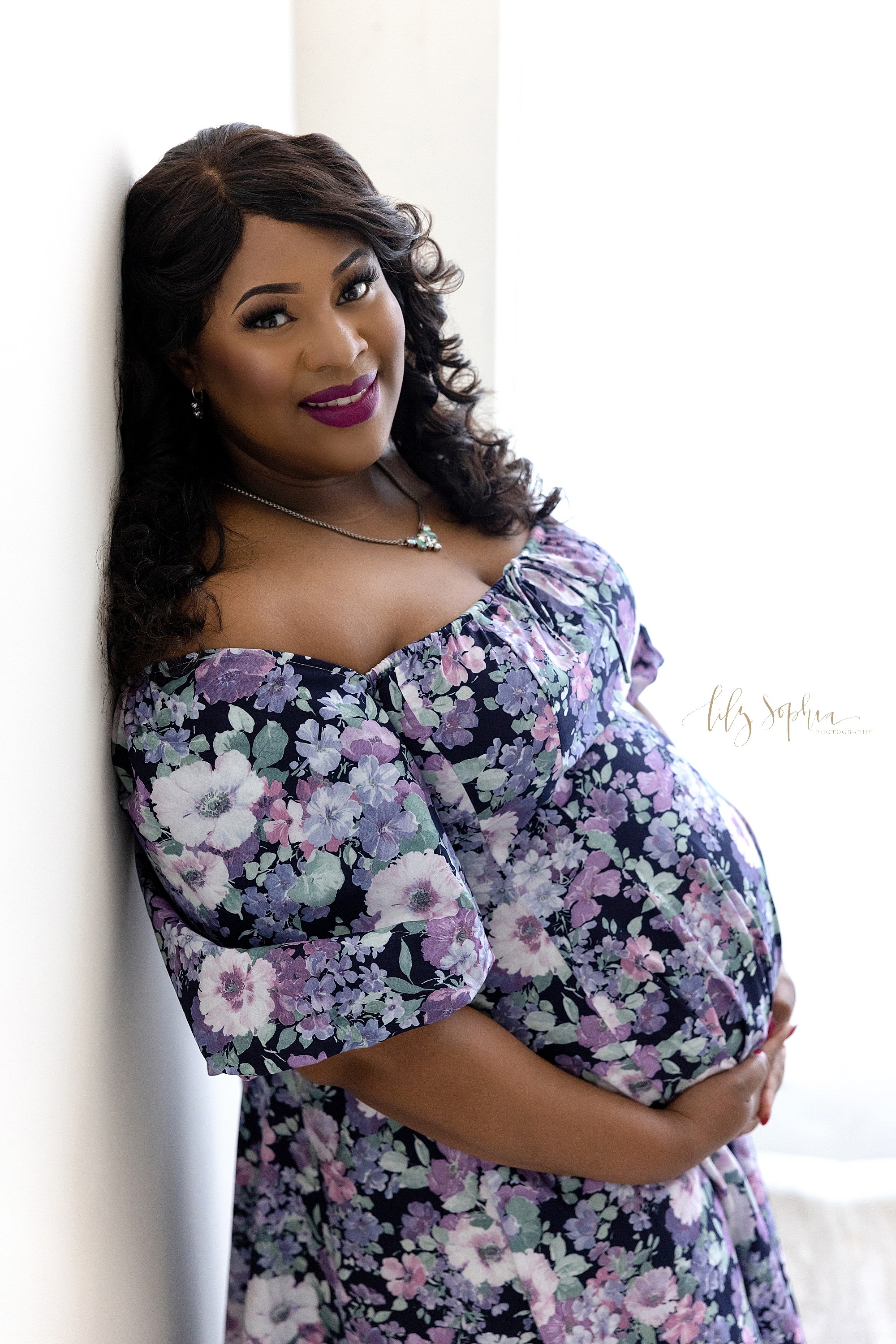  Maternity portrait of a pregnant African-American woman as she stands with her back against a wall next to a window streaming natural light as she holds the base of her belly taken near Poncey Highlands in Atlanta in a photography studio. 