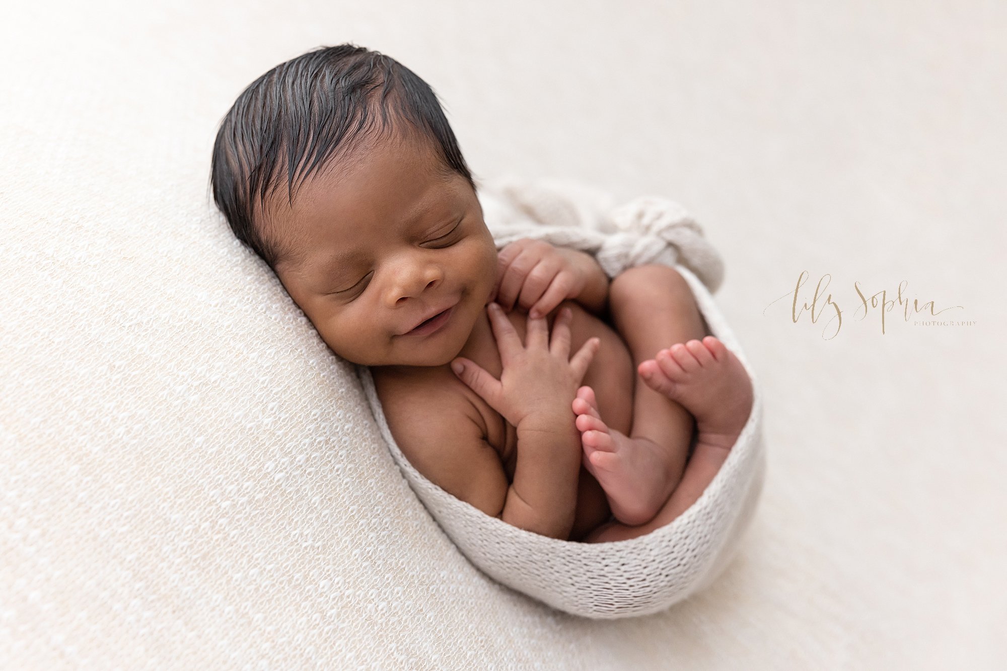  Newborn portrait of a sleeping African-American newborn baby boy cradled in a blanket as he smiles taken using natural light in a studio near Old Fourth Ward in Atlanta, Georgia. 