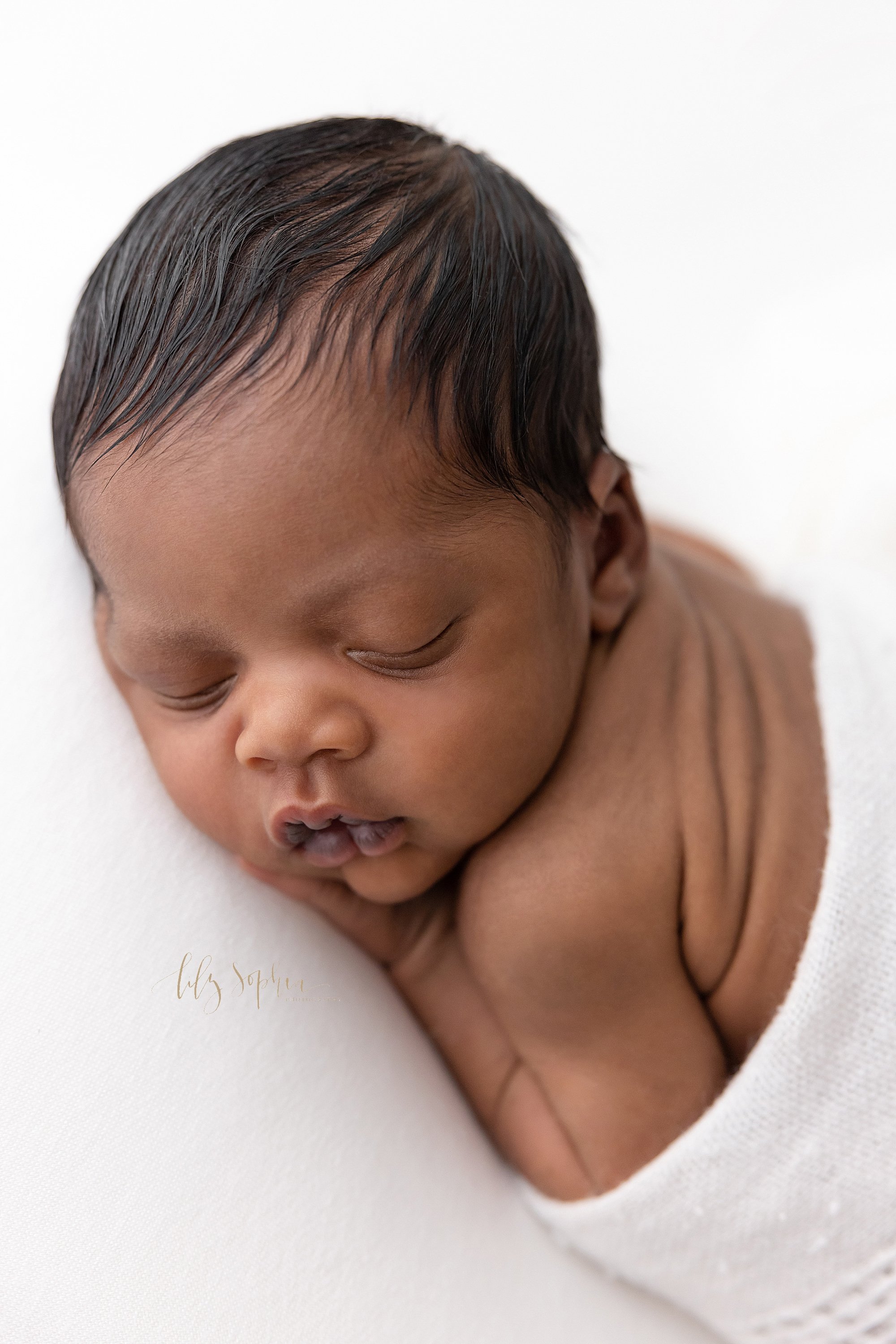  Close-up newborn portrait of an African-American newborn baby boy as he lies on his stomach with his hands under his head while turning his head to his left with his wrinkly skin on his back showing taken using natural light in a studio near Kirkwoo