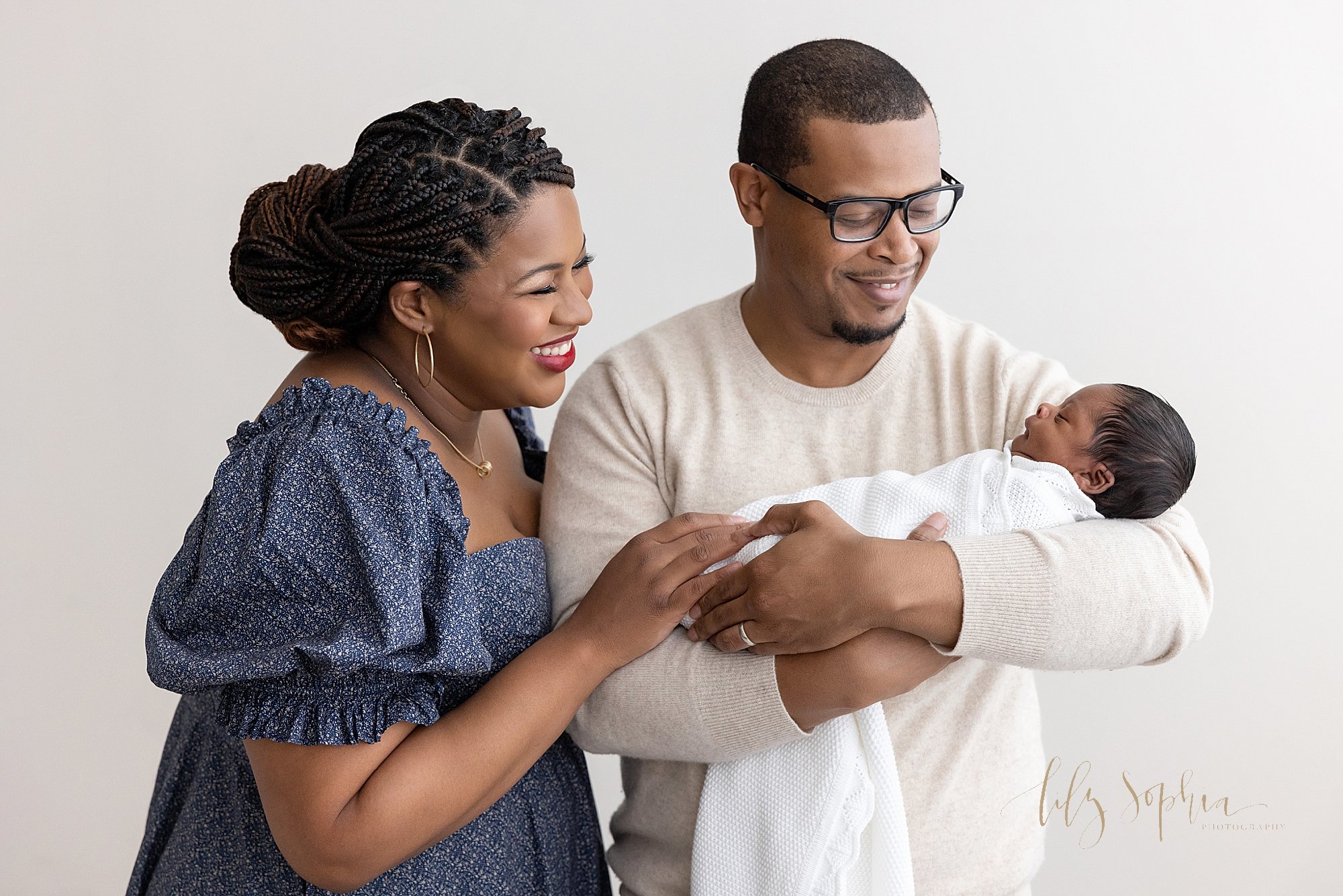  Family newborn photo session with an African-American father cradling his newborn son in his arms as mom stands to his right and smiles at their son taken in a studio near Oakhurst in Atlanta that uses natural light. 