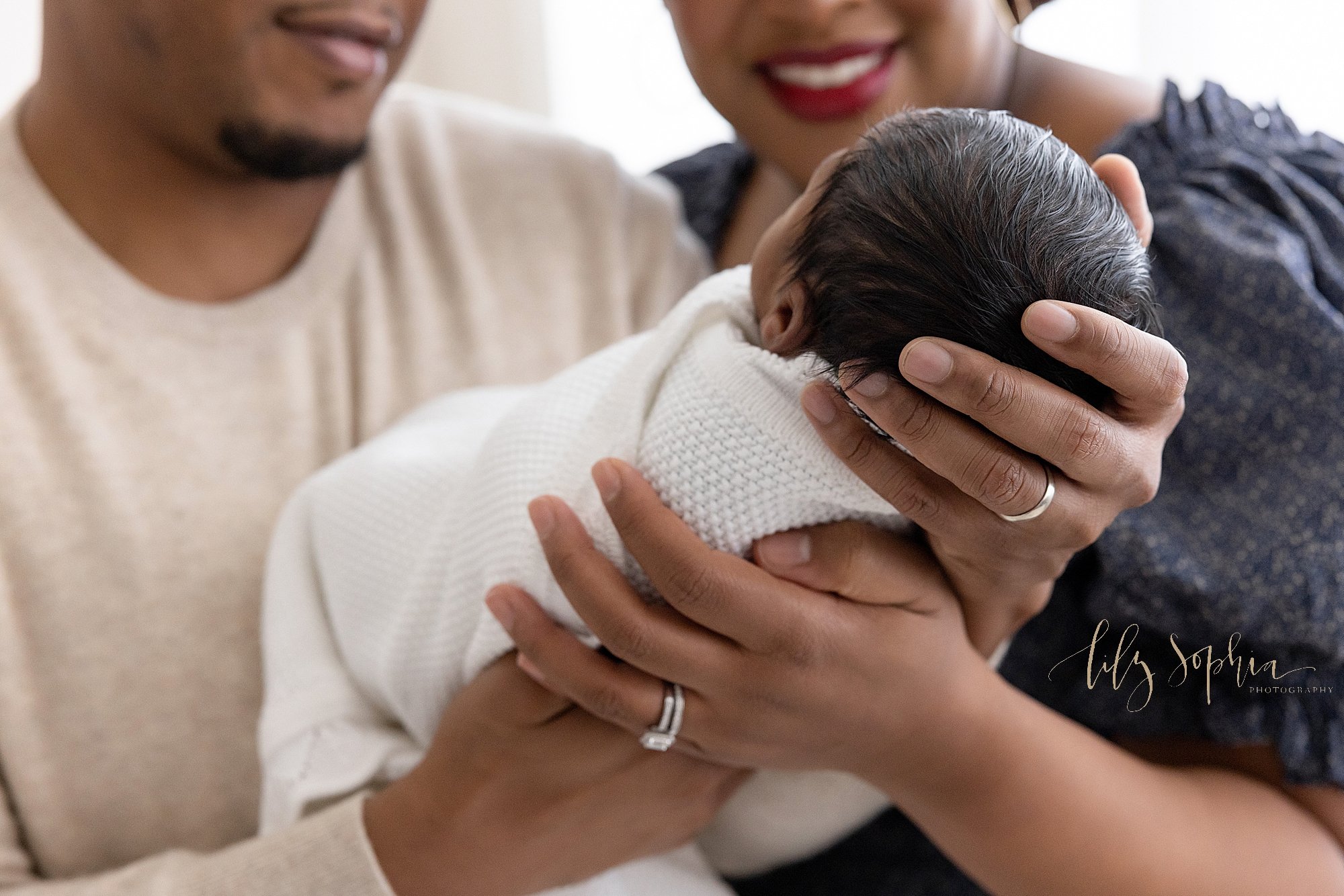  Newborn photo shoot of an African-American newborn baby boy’s head being held in his father’s hand as his mother stands to the right of his father and the two of them admire their son taken near Virginia Highlands in Atlanta in a natural light studi