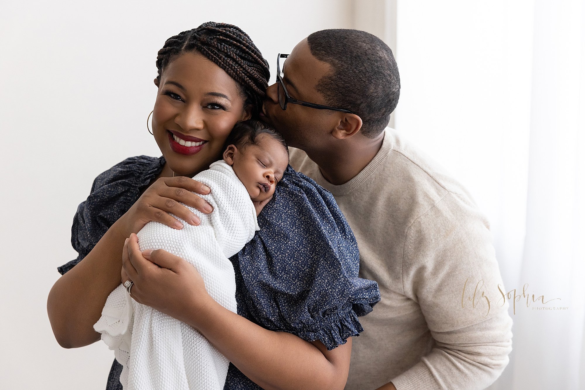  Newborn family photo of an African-American mother holding her peacefully sleeping newborn son on her shoulder as her husband kisses the left side of her head as he stands behind her and in front of a window streaming natural light in a studio near 