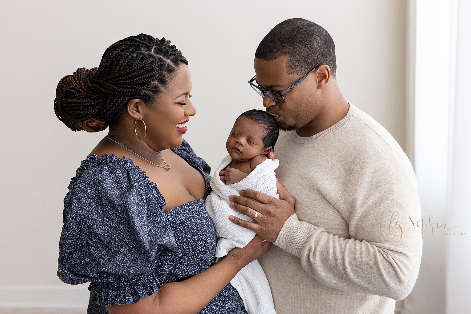  Family newborn portrait of an African-American mother holding her newborn baby son in front of her as her husband faces her and kisses the crown of her son’s head taken near Ansley Park in Atlanta in a studio using natural light. 