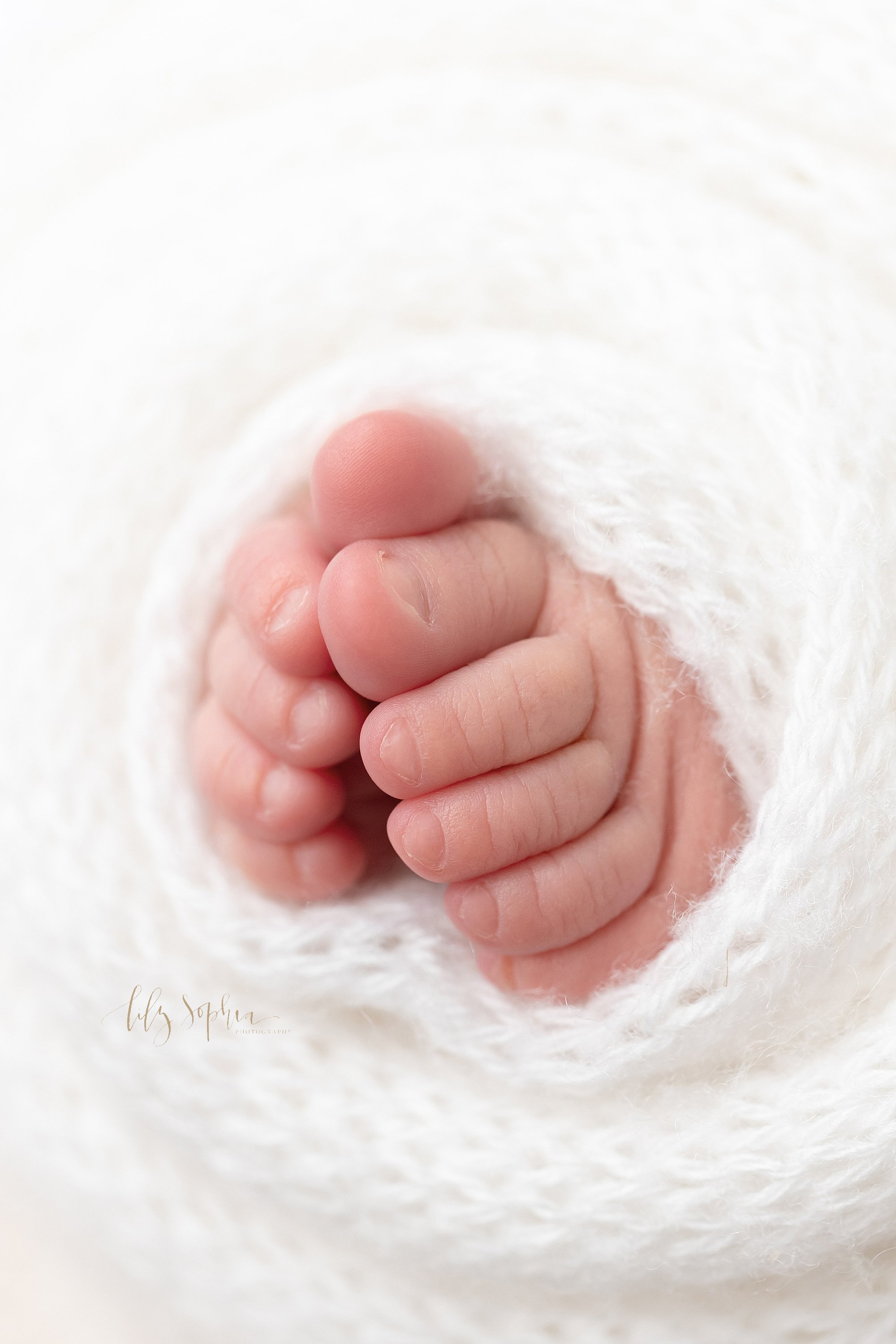  Newborn picture of a newborn baby girl’s tiny toes sticking out from a soft white blanket taken in a natural light studio near Buckhead in Atlanta. 