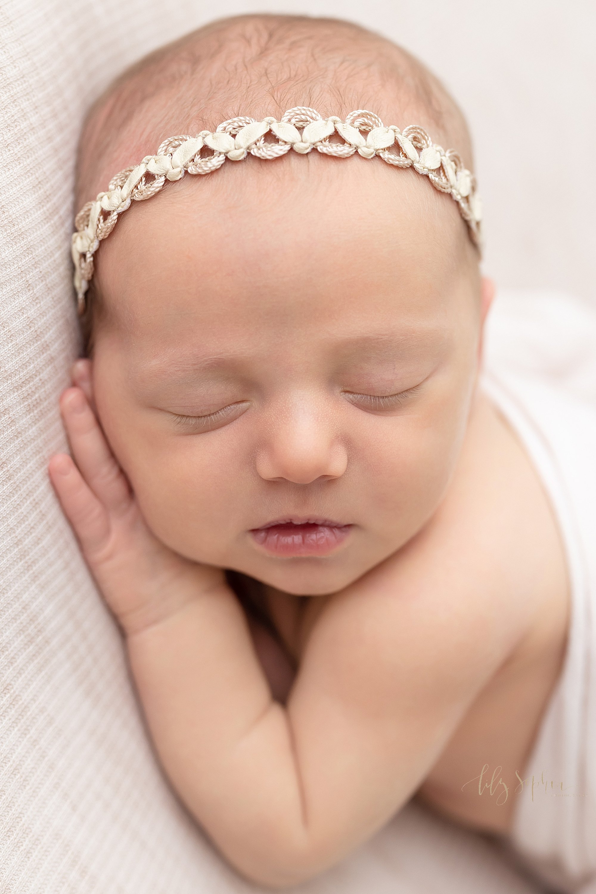  Close-up newborn portrait of a newborn baby girl lying on her stomach with her head turned to her right as her cheek rests on her right hand featuring her milky lips, her button nose, and her wispy eye lashes taken near Midtown in Atlanta in a studi