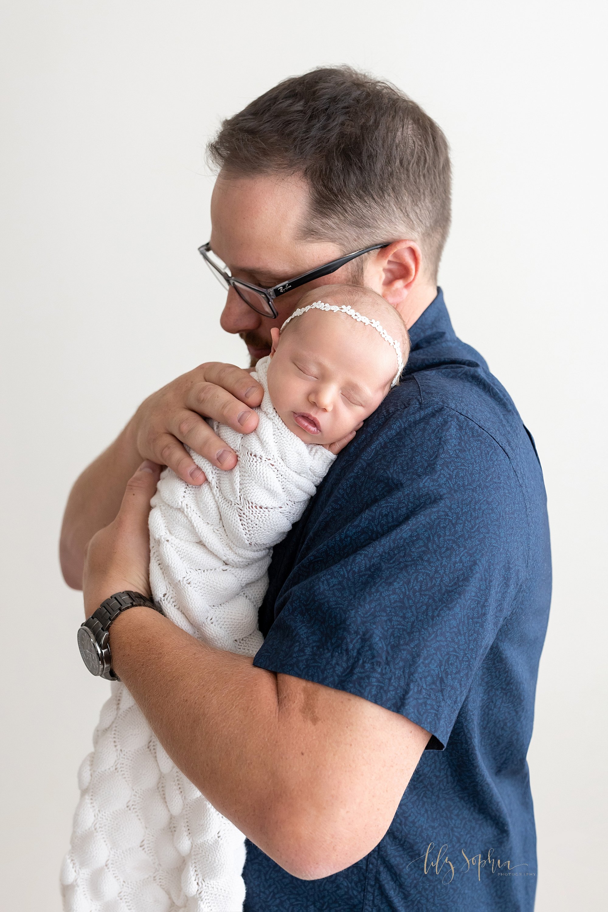  Newborn portrait of a father holding his newborn baby girl on his shoulder as she peacefully sleeps taken in front of a window streaming natural light in a studio located near Morningside in Atlanta, Georgia. 