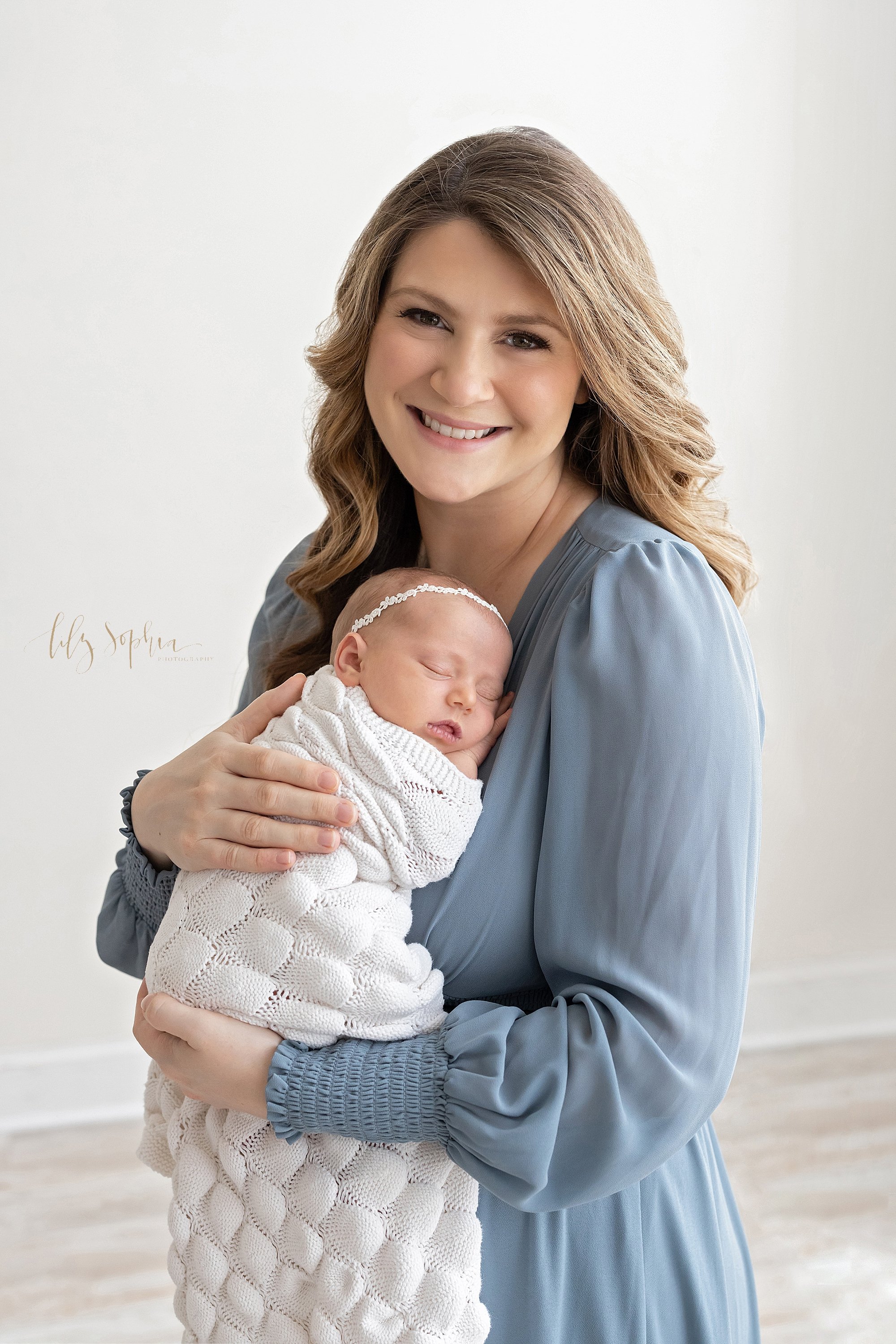  Newborn portrait of a happy mother holding her sleeping newborn baby daughter against her chest as she stands next to a window streaming natural light in a photography studio near Poncey Highlands in Atlanta. 