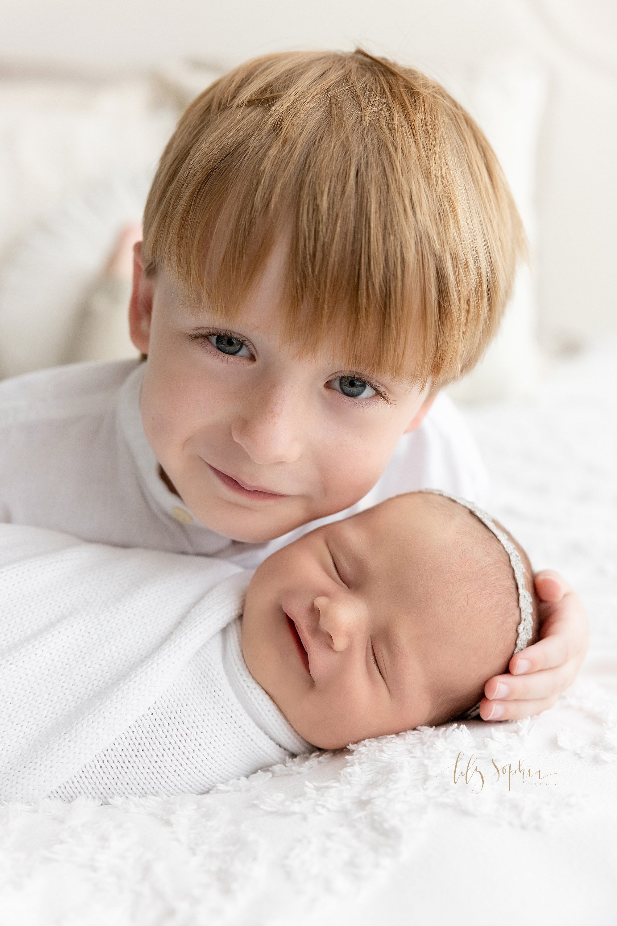  Sibling newborn portrait of a young boy lying on his stomach on a bed with his sleeping newborn baby sister in front of him as they both smile while the boy places his hand on her head taken using natural light in a photography studio in Ponce City 