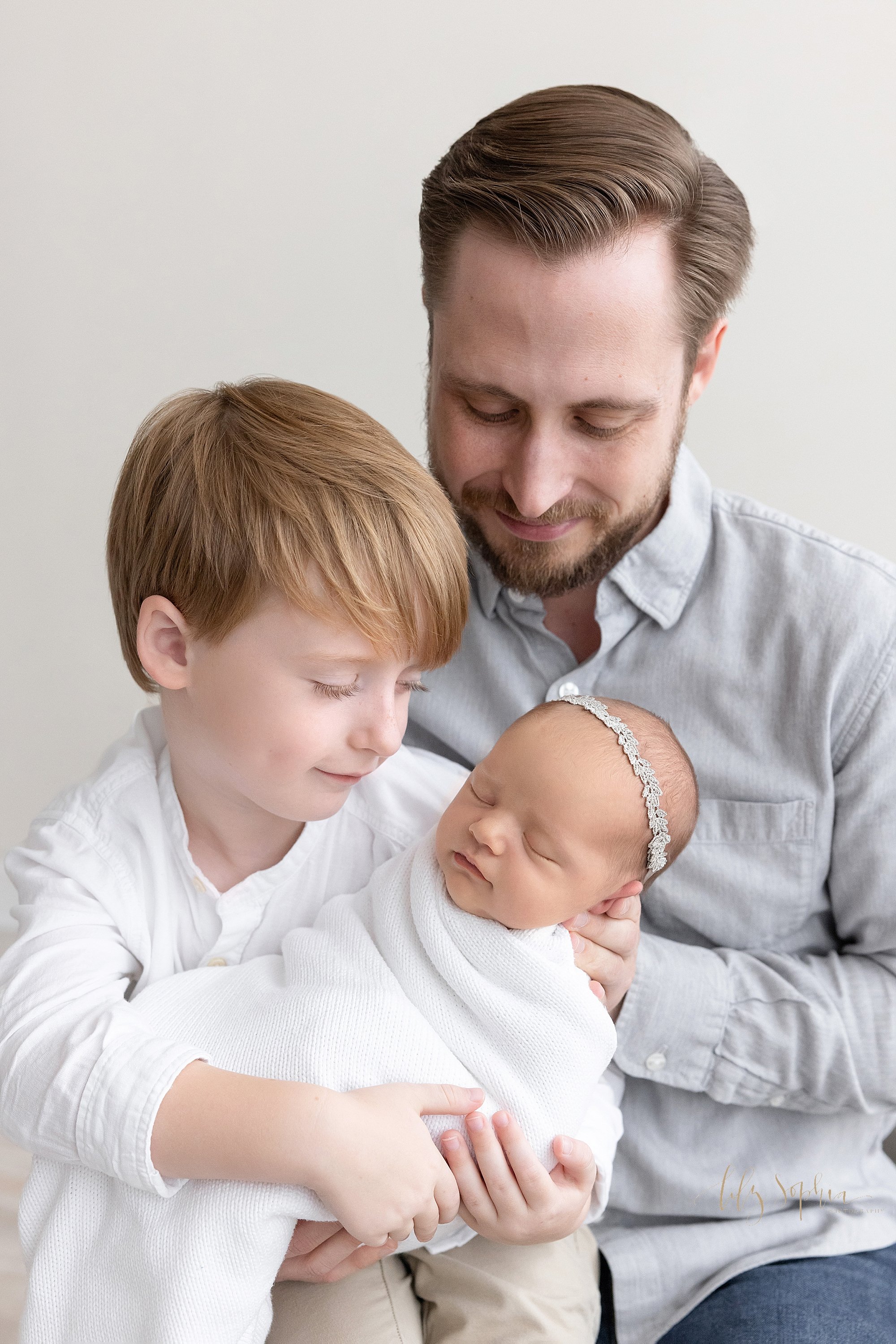  Family picture of a young son holding his newborn baby sister in his arms as he sits on his father’s right knee and his father looks on taken in natural light near Virginia Highlands in Atlanta in a photography studio. 