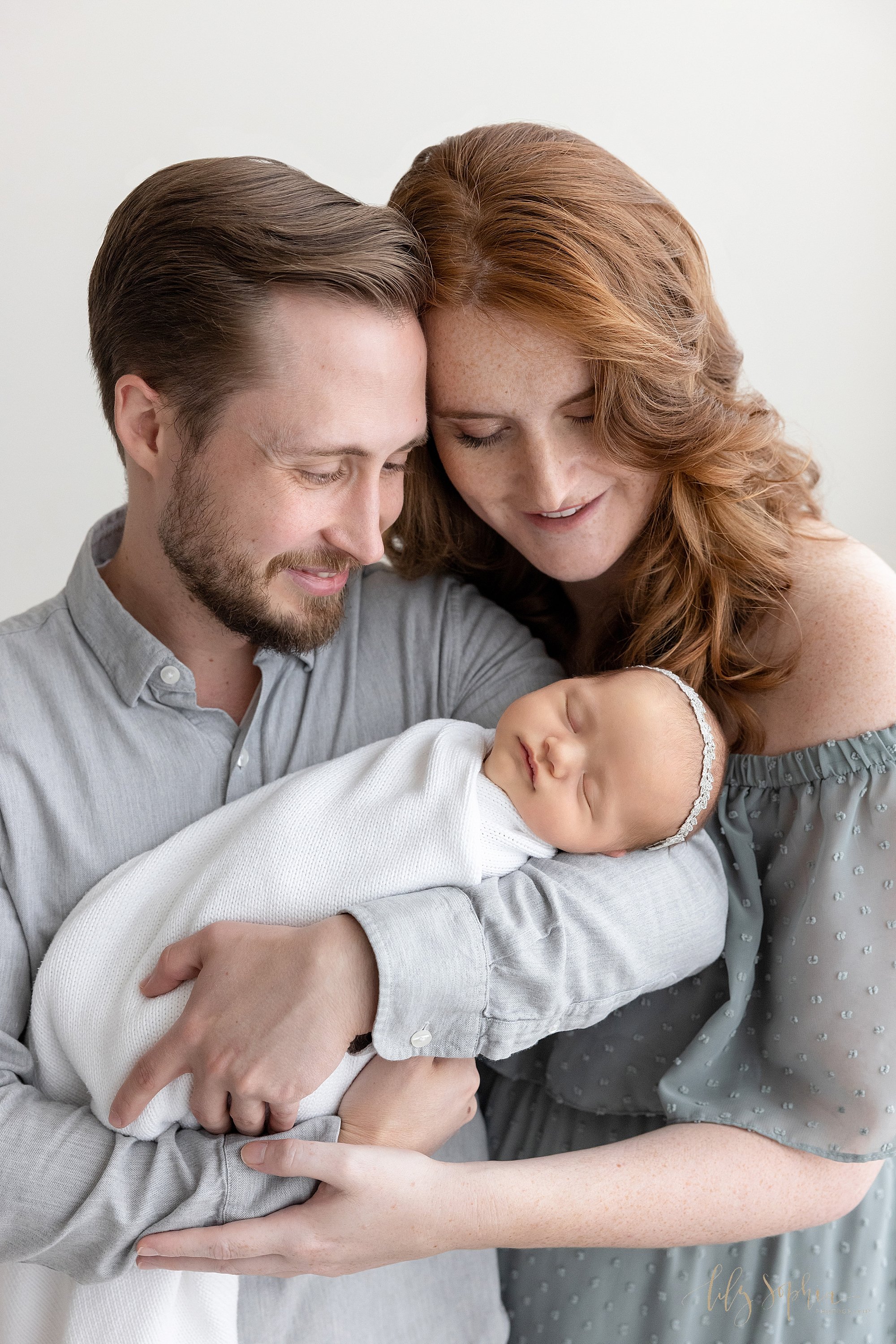  Newborn portrait of a father cradling his newborn baby girl in his arms as his wife stands next to him on his left and they place their heads together and admire their peacefully sleeping daughter taken near Oakhurst in Atlanta in a photography stud