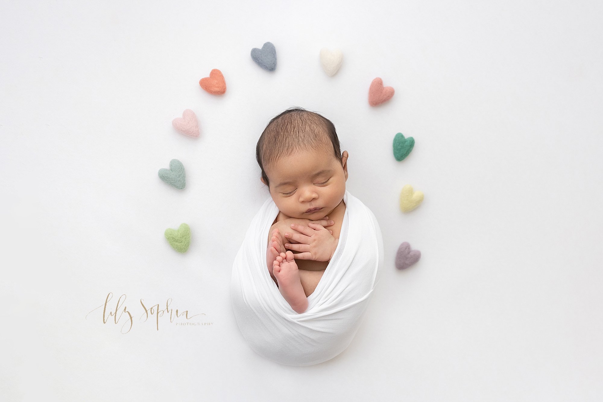  Newborn portrait of an Indian newborn baby boy cradled in a stretchy swaddle with  different colored hearts arching around him taken using natural light near Brookhaven in a photo studio. 