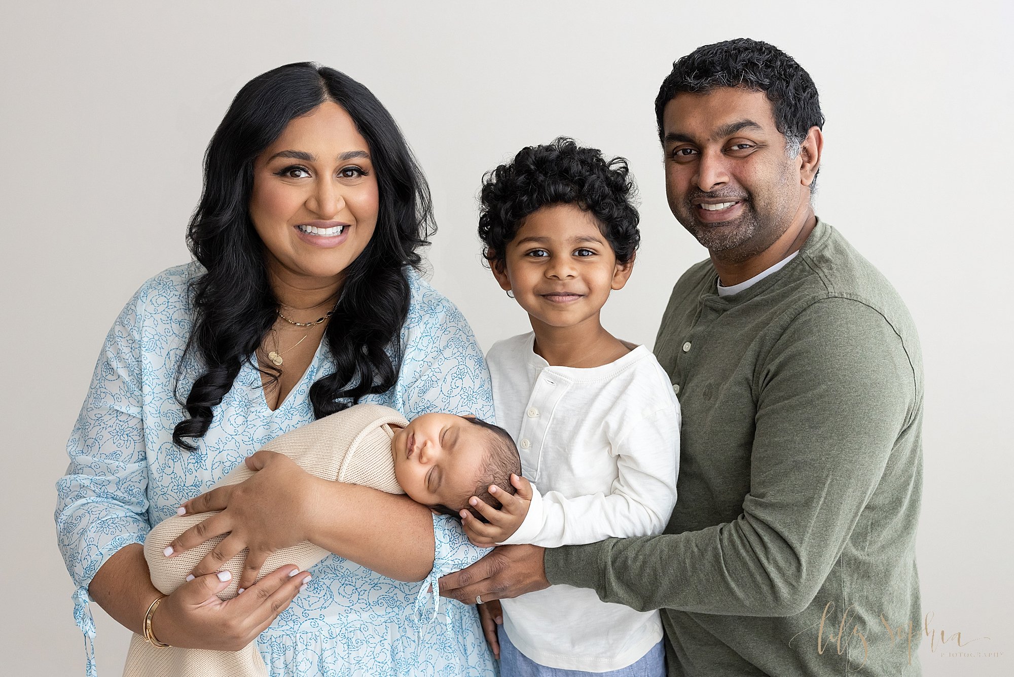  Family newborn photo of an Indian mother cradling her sleeping newborn son in her arms as her young son stands to her left and places his hand on his brother’s head and his father stands next to him with his arm supporting his wife taken near Smyrna