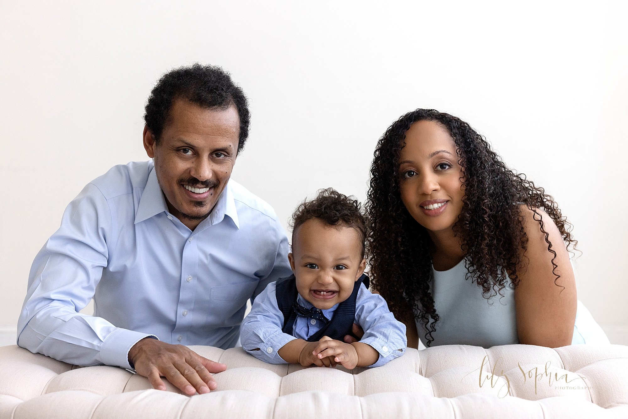  Family portrait of a father and mother with their one year old son between them as the parents kneel and their son stands behind a tufted bench in a natural light studio near Cummings in Atlanta, Georgia. 