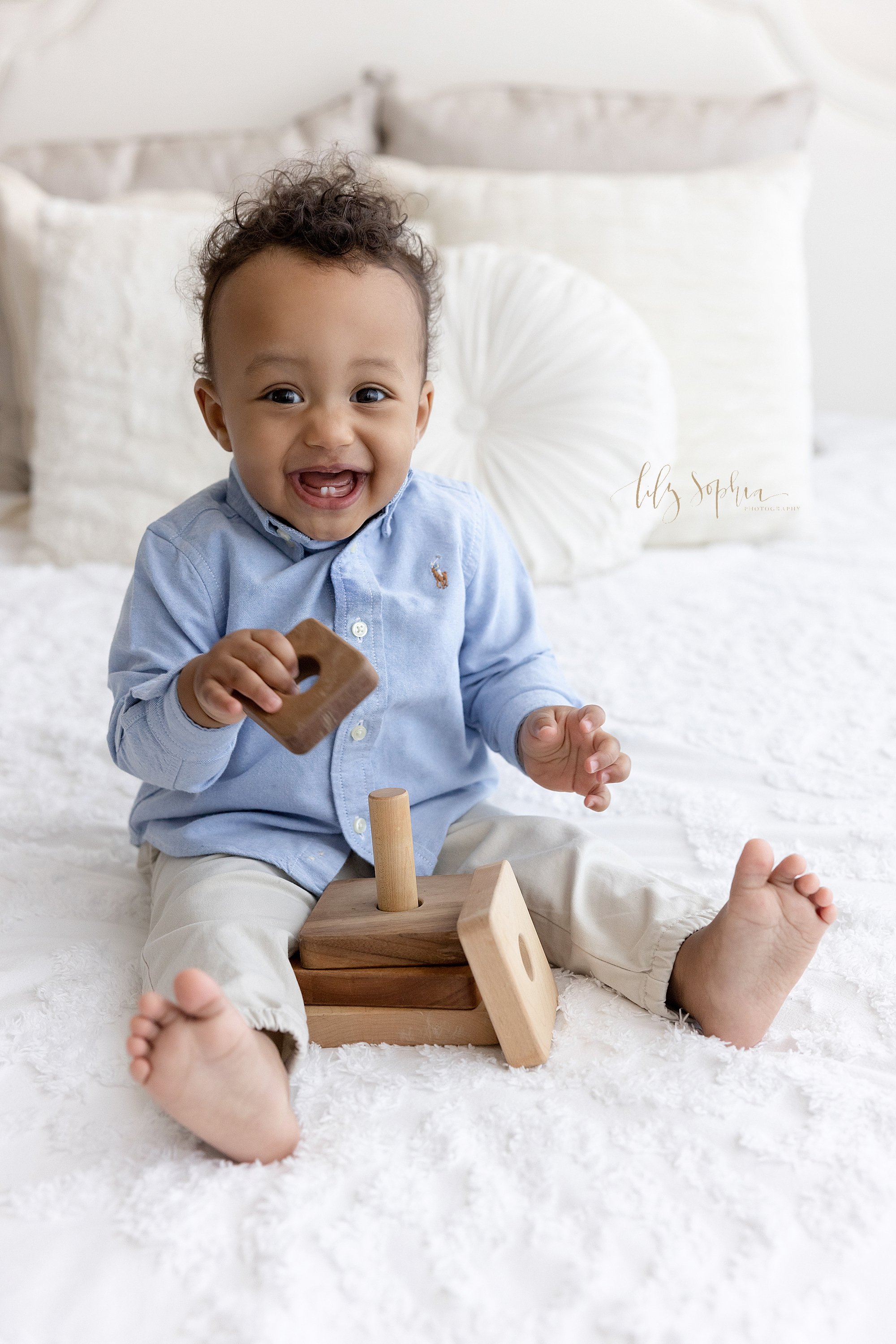 First birthday photo session of an African-American little boy as he sits atop a bed playing with a wooden stacking toy and laughs taken in a natural light photography studio near Smyrna in Atlanta. 