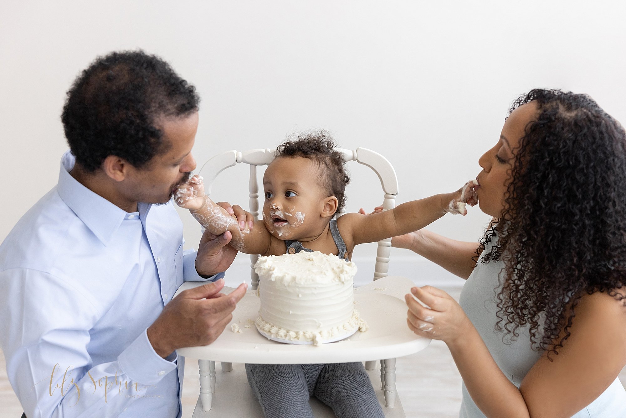  First birthday smash cake session of a one year old little boy sitting in an antique high chair with icing covering his face as he feeds his father on his right and his mother on his left taken in a studio near Oakhurst in Atlanta that uses natural 