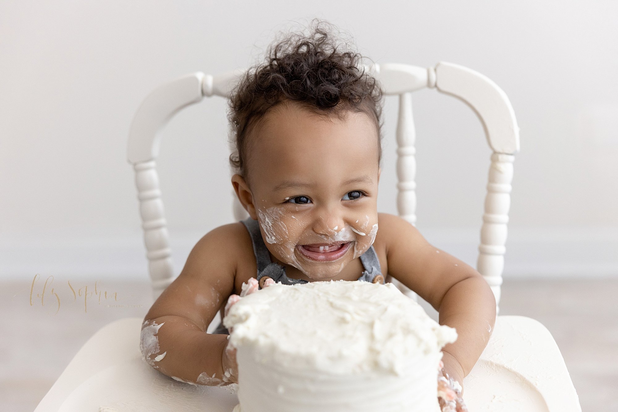  First birthday smash cake picture of an African-American one year old boy as he sits smiling with icing on his face in an antique high chair with his smash cake in front of him taken next to a window streaming natural light in a studio near Decatur 