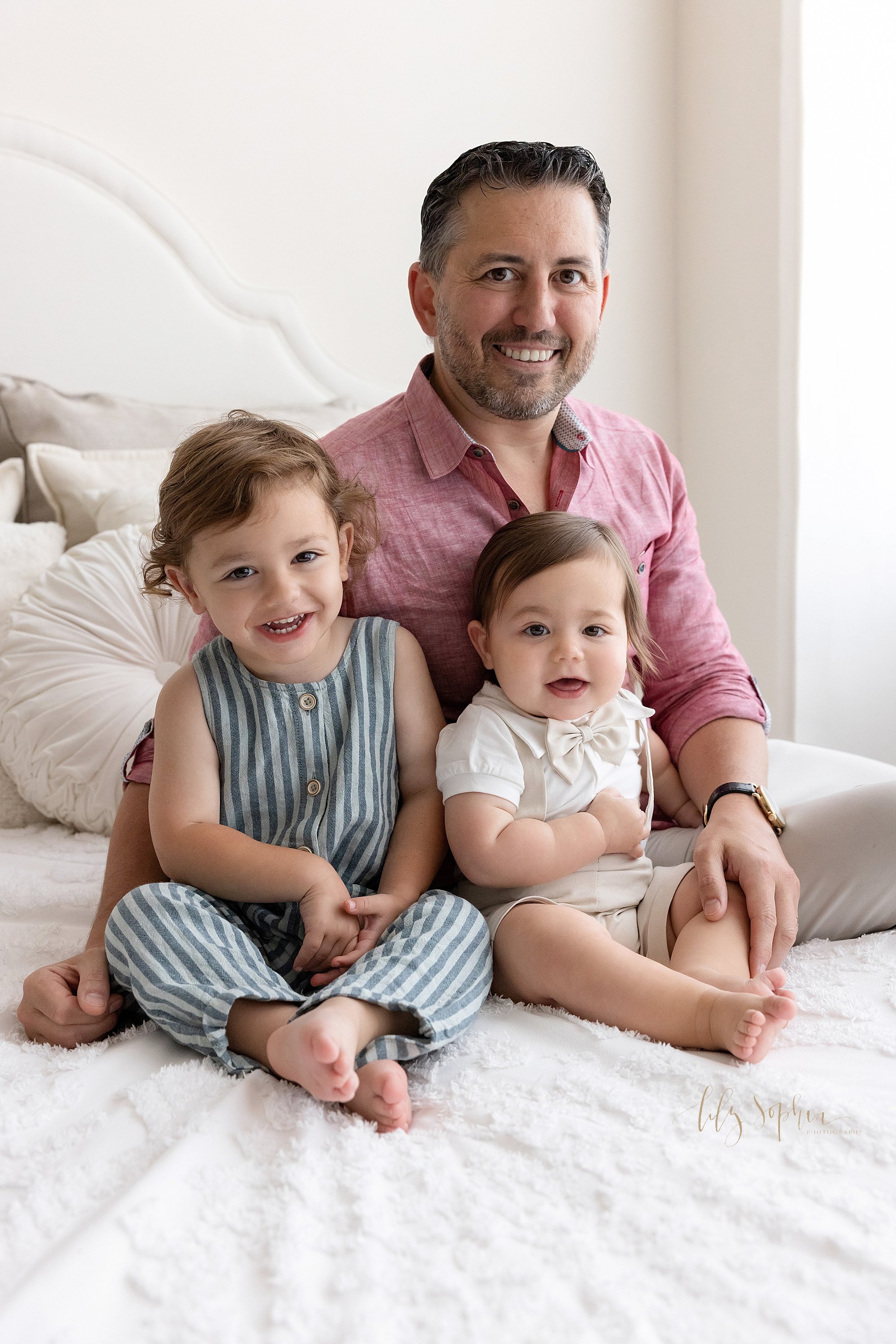  Family portrait of a father sitting on a bed next to a window streaming natural light with his young daughter sitting in front of him to his right and his one year old son sitting next to his sister taken near Ansley Park in Atlanta in a photography