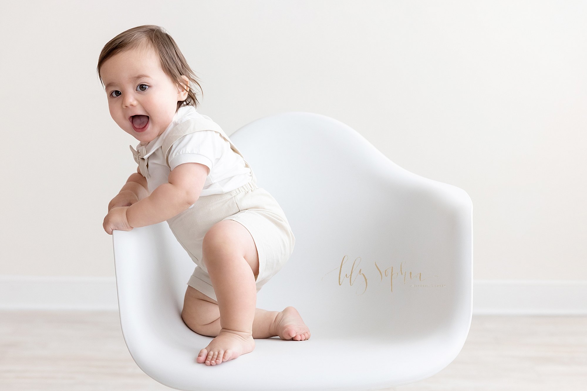  First birthday photo session of a rambunctious one year old boy as he tries to stand up in a white molded chair taken in a natural light photography studio located near Poncey Highlands in Atlanta. 
