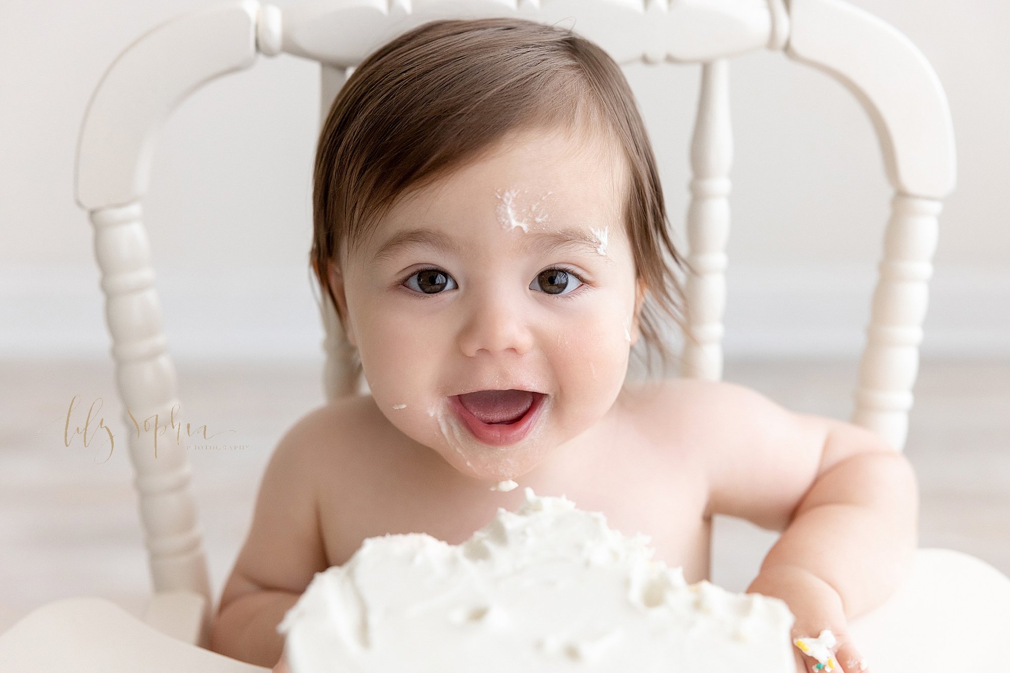  First birthday smash cake photo session of a one year old boy with icing on his face as he sits in an antique high chair with his smash cake on the tray taken using natural light in a photography studio near Morningside in Atlanta. 