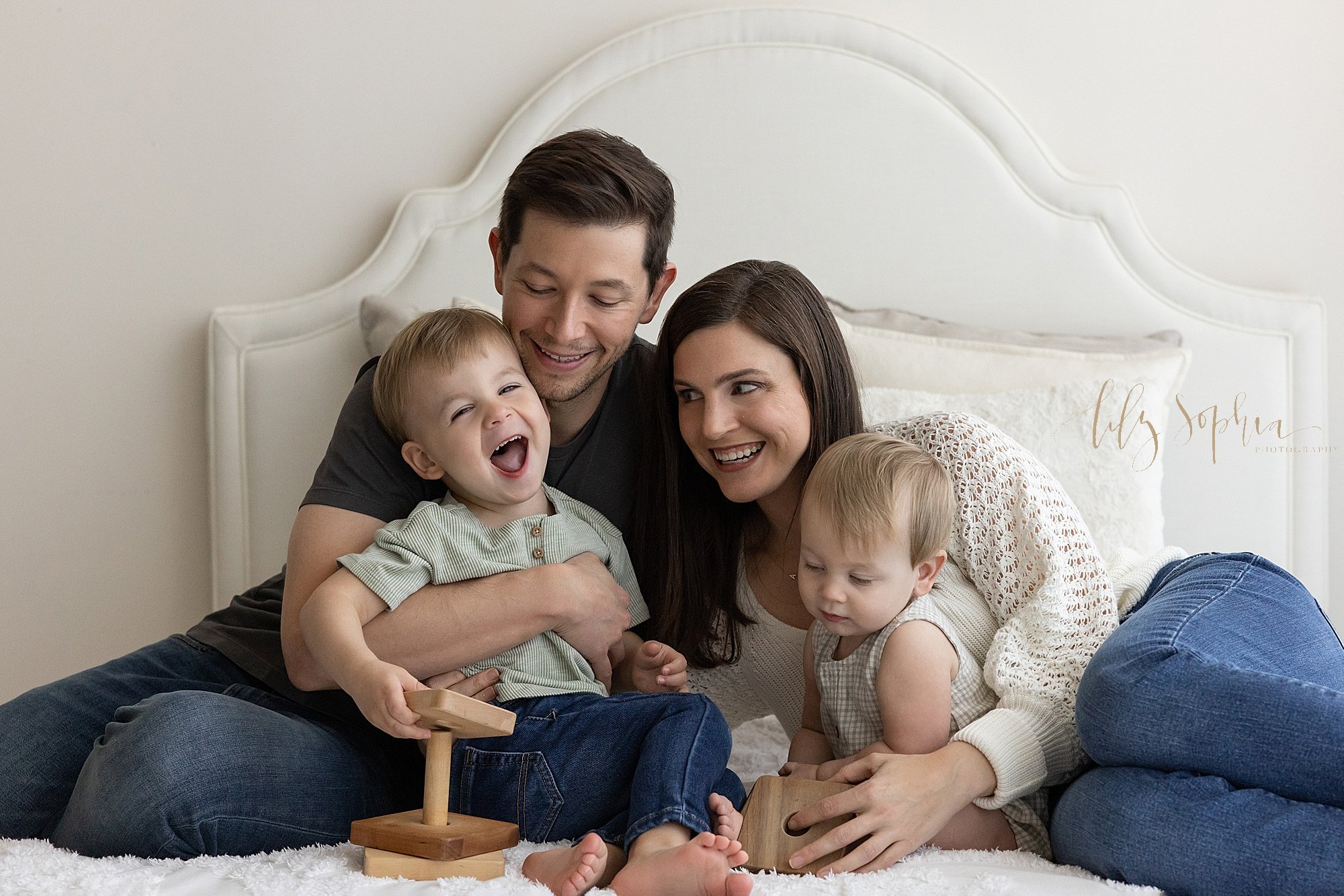  Fun family photo of a father holding his laughing toddler son in front of him on a bed as the son plays with a wooden stacking toy and his wife leans on her right arm while lying on the bed next to them with their one year old son in front of her ta
