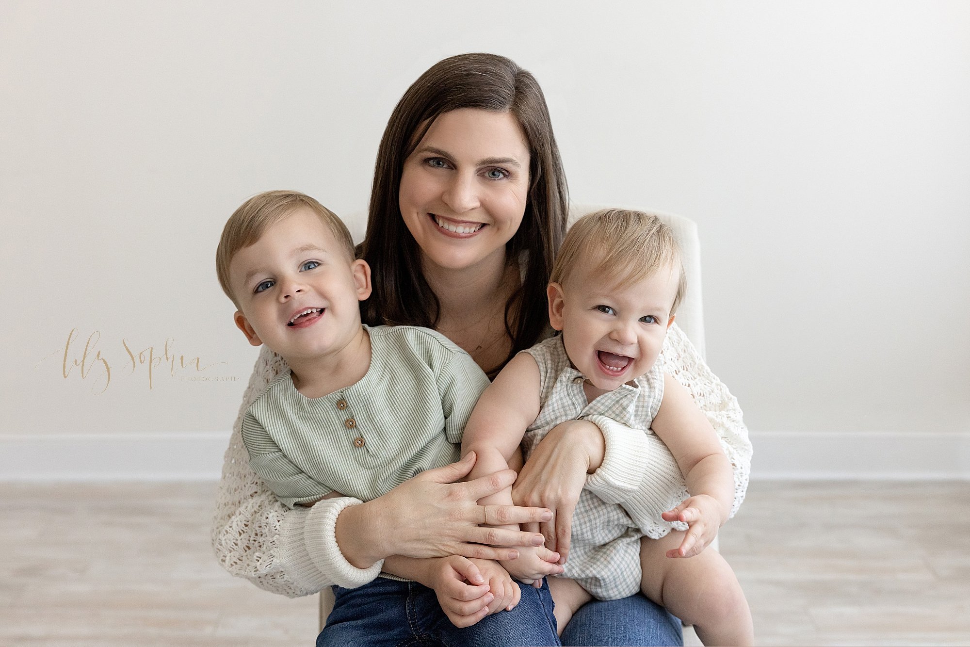  Family photo of a mom sitting in a photography studio with her toddler son on her right knee and her one year old son on her left knee as they all laugh and smile taken near Alpharetta in Atlanta in a natural light. 