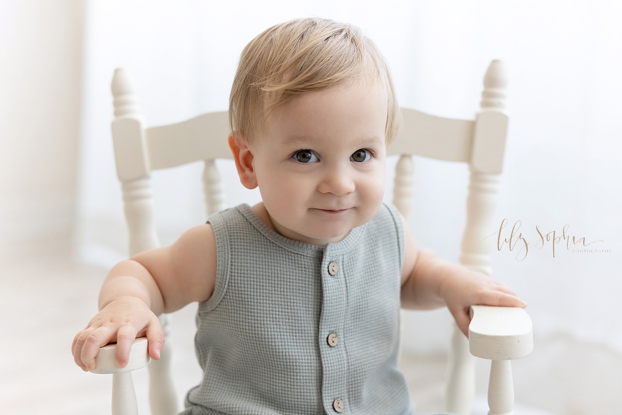  First birthday picture of a one year old baby boy in front of a window streaming natural light sitting in a white rocking chair taken near Brookhaven in Atlanta in a photography studio. 