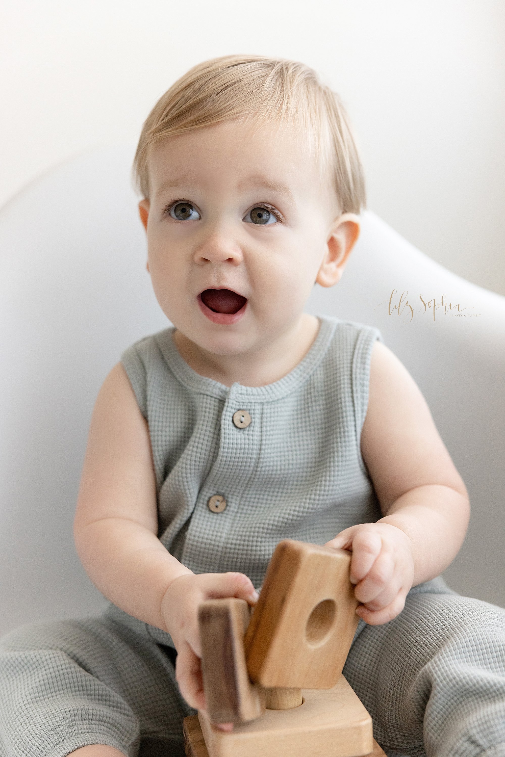  Close-up first birthday portrait of a one year old baby boy as he sits in a white molded chair, looks up with his big blue eyes and plays with a wooden stacking toy taken near Buckhead in Atlanta in a photography studio that uses natural light. 