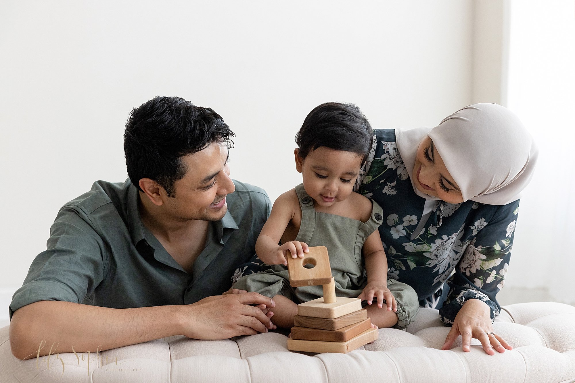  Family photo of a one year old little boy as he sits between his parents on a tufted bench and plays with a wooden stacking toy as his parents watch him taken near Morningside in Atlanta in a studio with a window streaming natural light. 