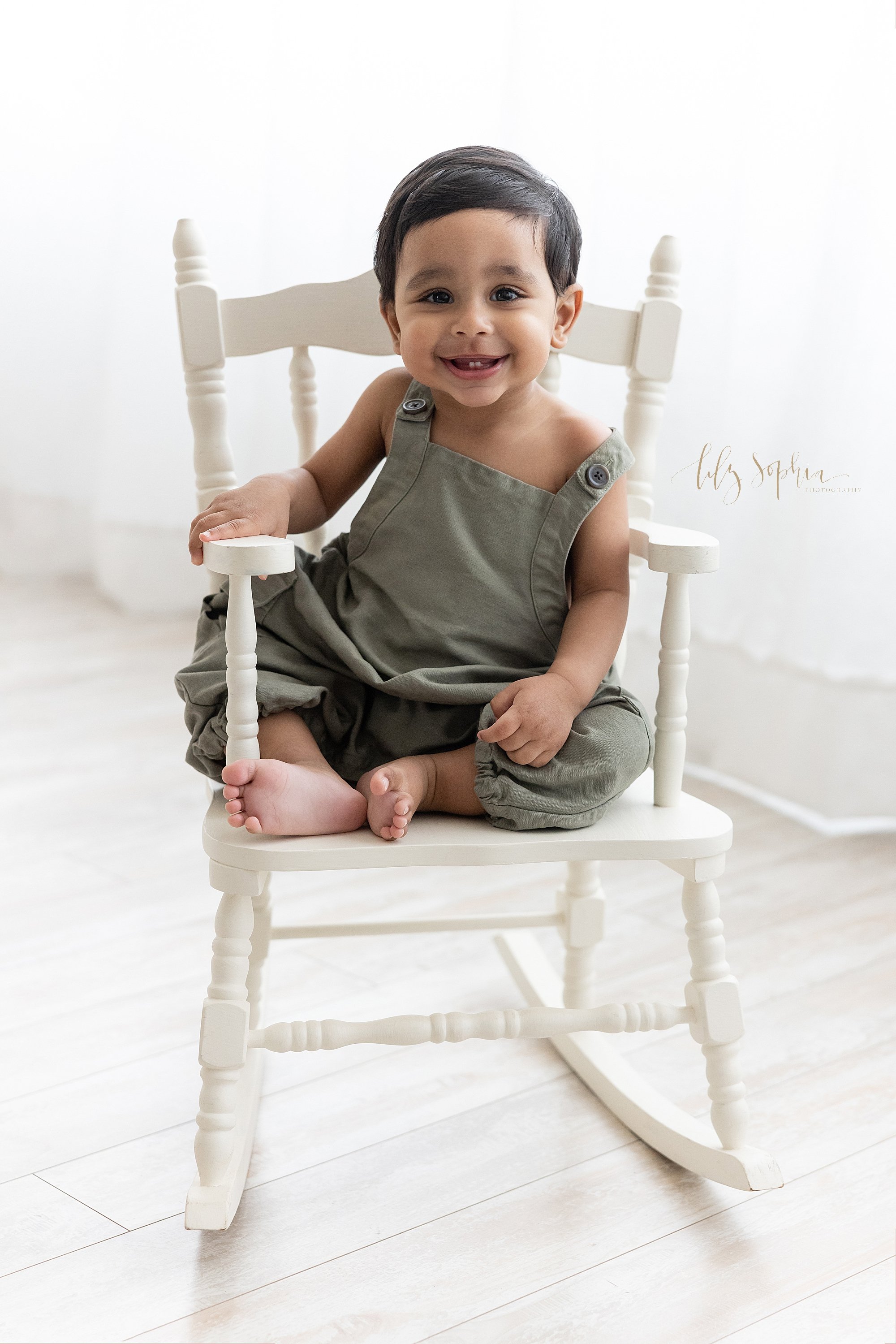  First birthday photo shoot of a one year old little boy as he sits in front of a window streaming natural light in a rocking chair smiling to show his tiny bottom baby teeth in a photography studio near Buckhead in Atlanta, Georgia. 