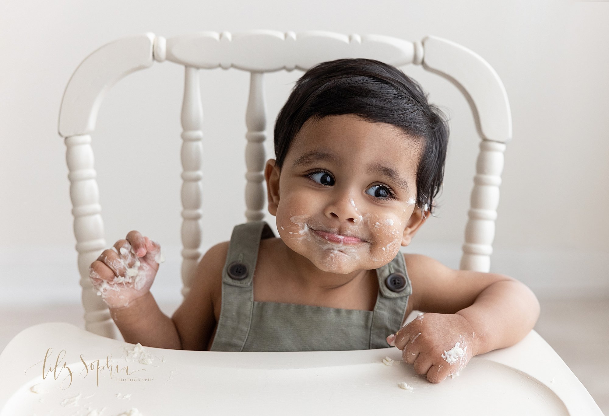  First birthday smash cake photo session with a one year old little boy sitting in an antique high chair with icing covering his face and hands as he smirks taken near Midtown in Atlanta in a studio that uses natural light. 