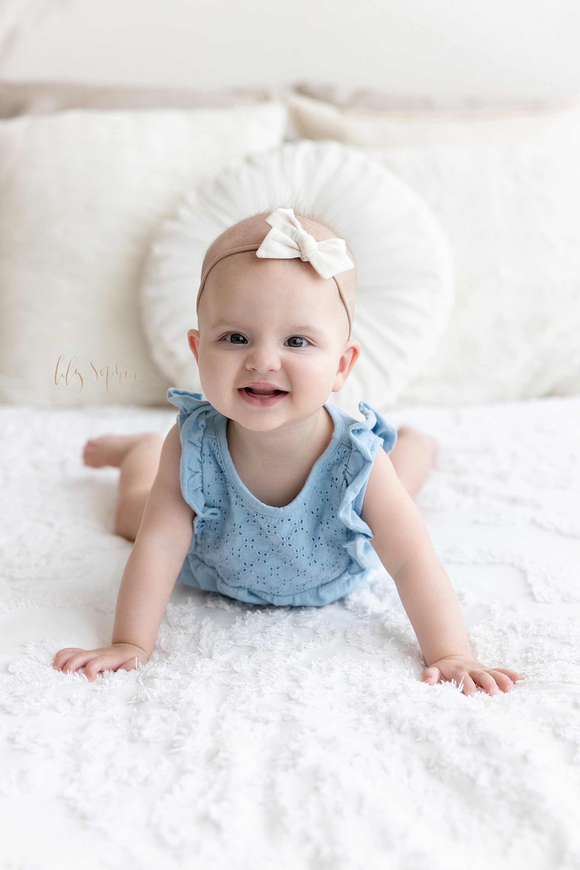  Milestone photo of a baby girl as she lies on her stomach on a bed and pushes herself up with her arms while smiling her toothless grin taken next to a window streaming natural light in a photography studio near Midtown in Atlanta. 
